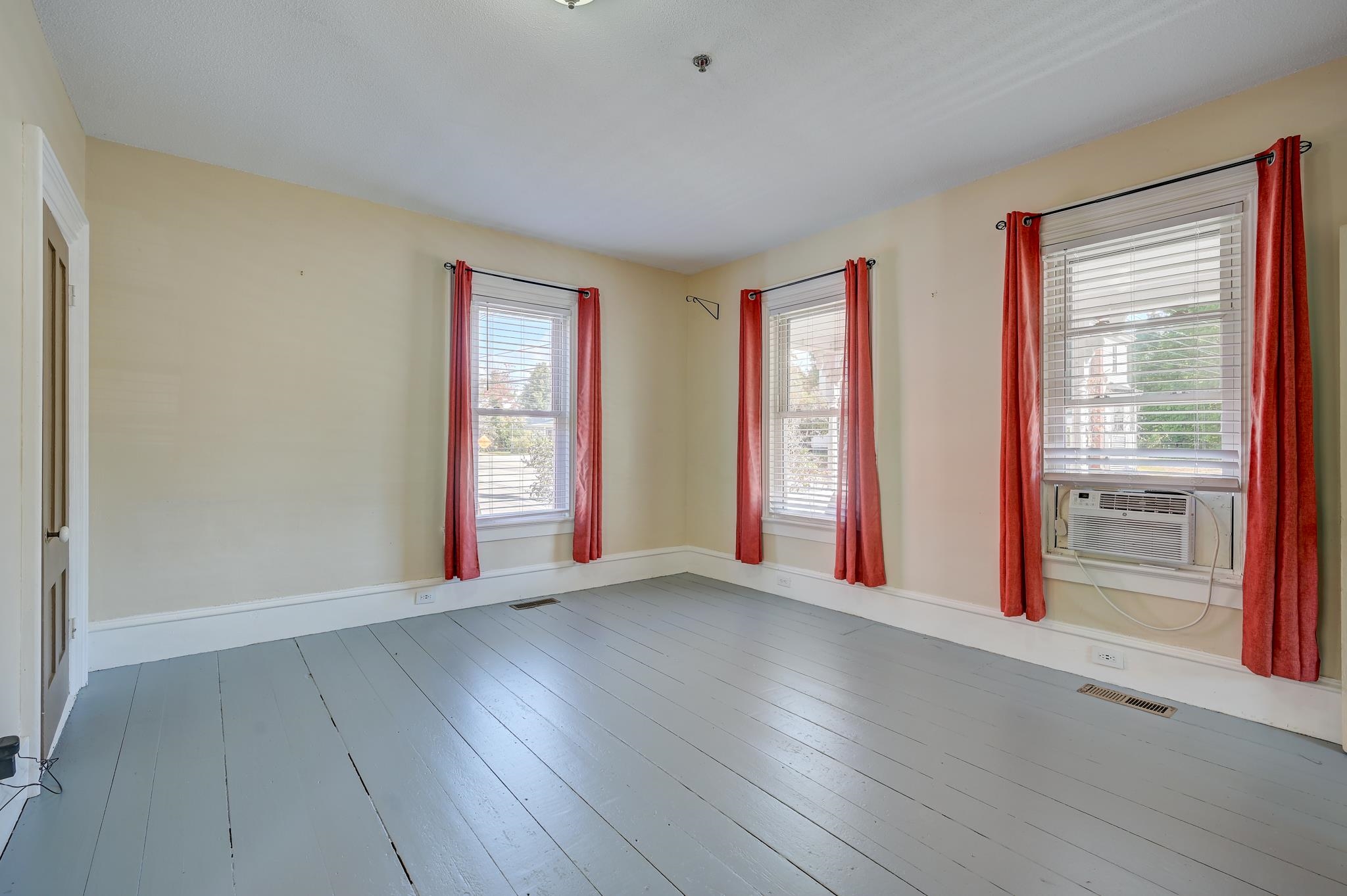 Empty room with gray plank floors, three curtained windows, and a window air conditioner.