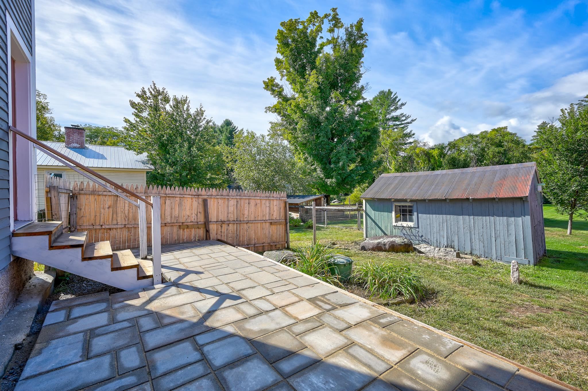 Paved patio and wooden steps beside a fenced backyard with a blue-gray shed and mature trees.