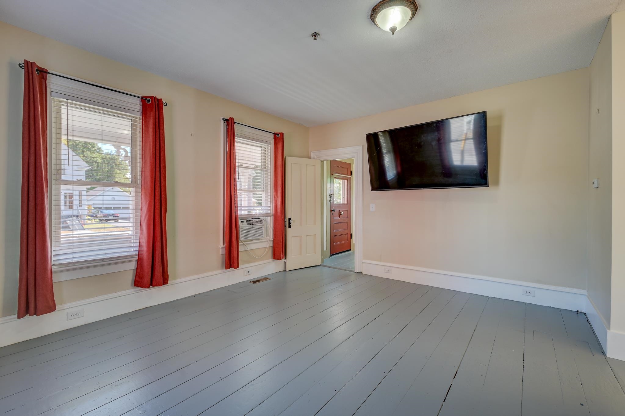 Empty room with gray plank floors, red window curtains, and a wall-mounted television.