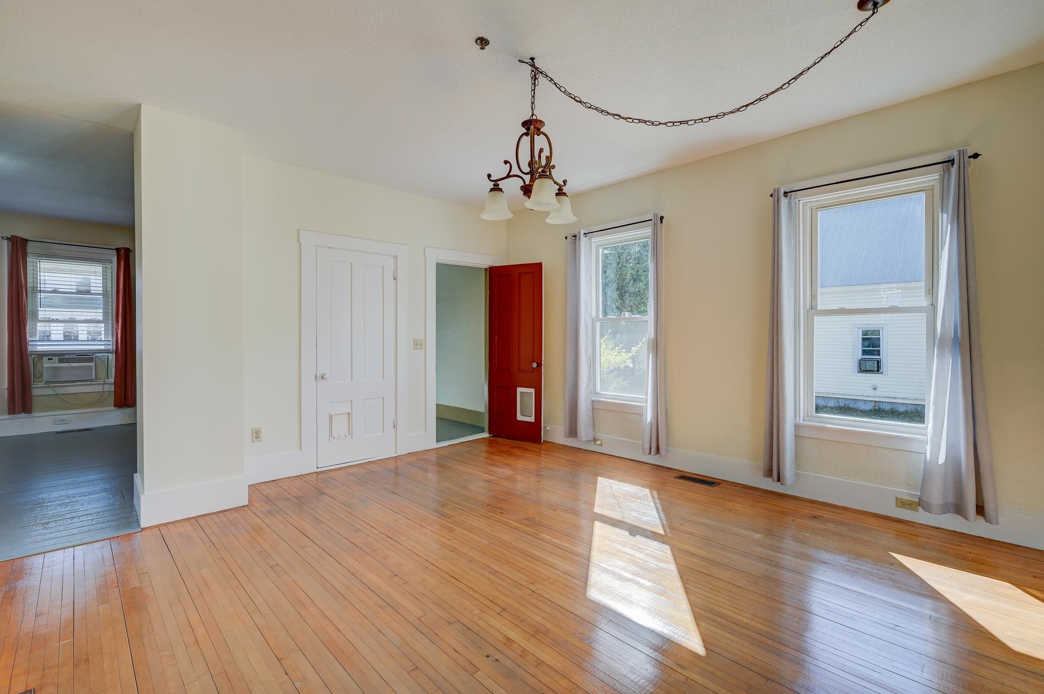 Unfurnished room with polished hardwood floors, a chandelier, two windows, and a red door with a pet door.