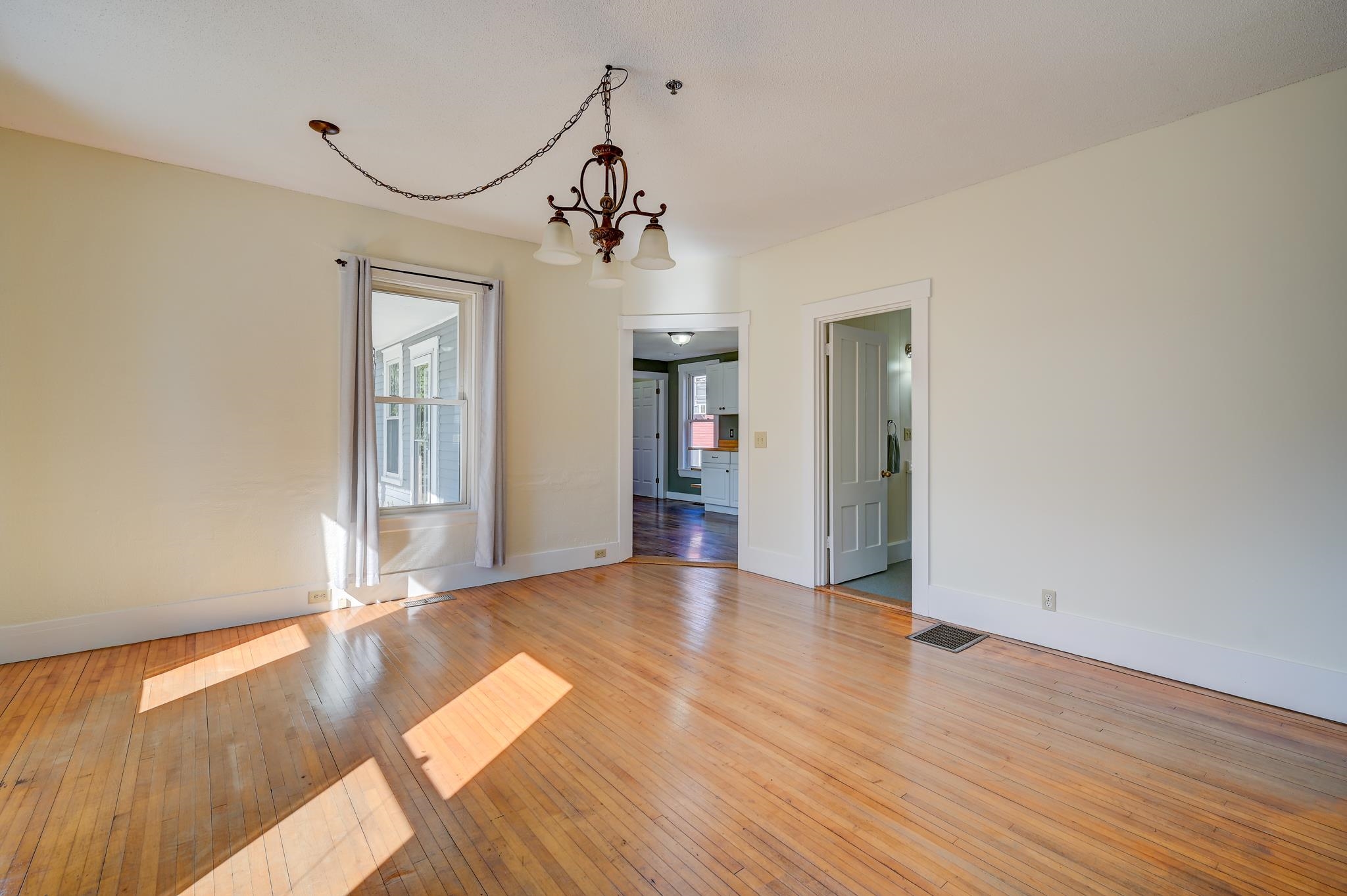 Empty room with polished hardwood floors, a chain-hung chandelier, and sunlight across the floor.