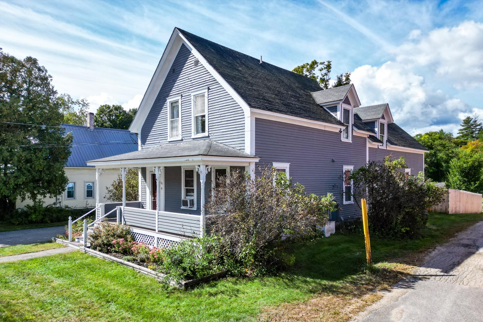 Gray clapboard house with a covered porch, dormer windows, and flower beds along the front.