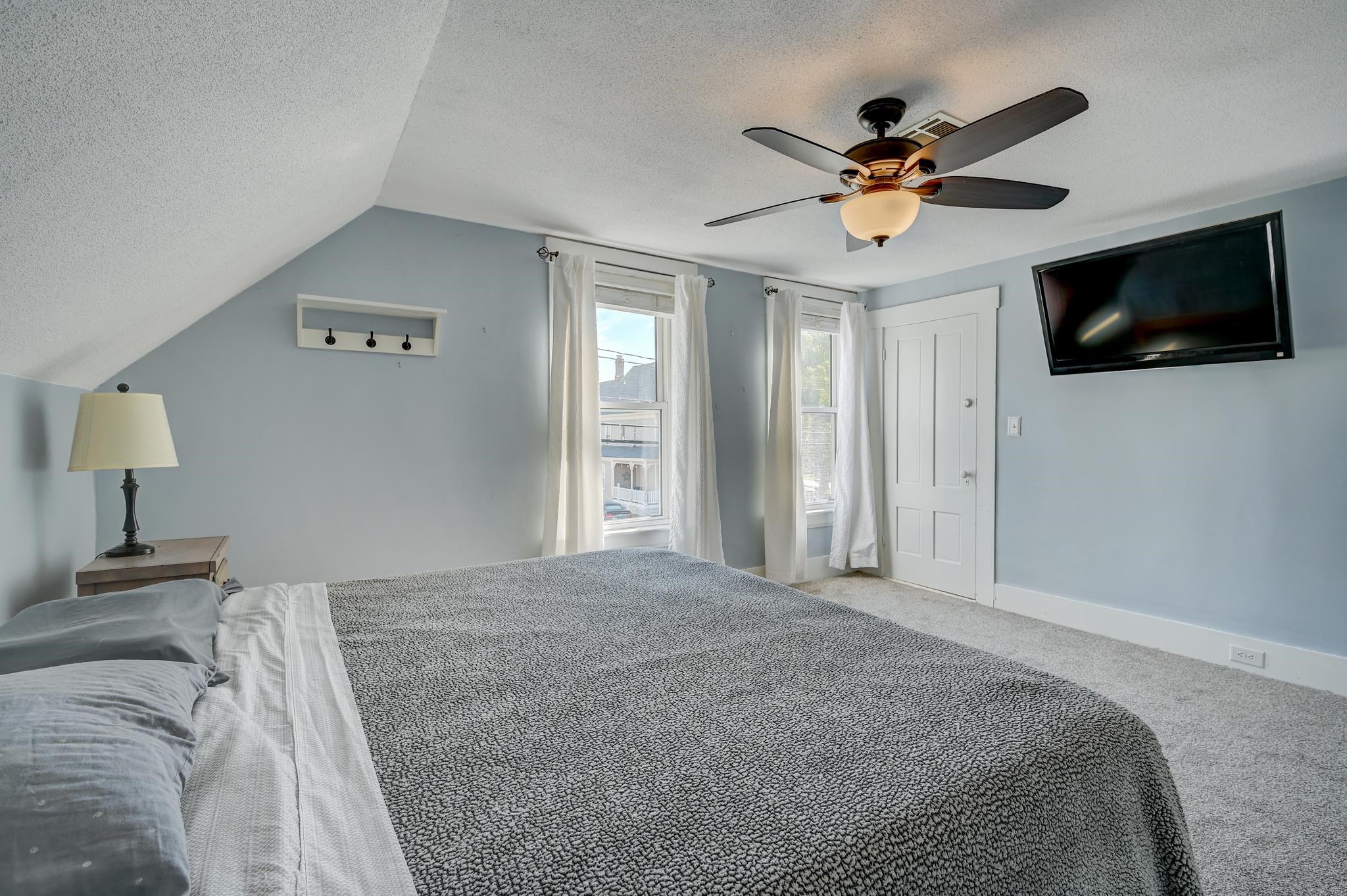 Furnished bedroom with gray patterned bedding, two curtained windows, and a wall-mounted television.