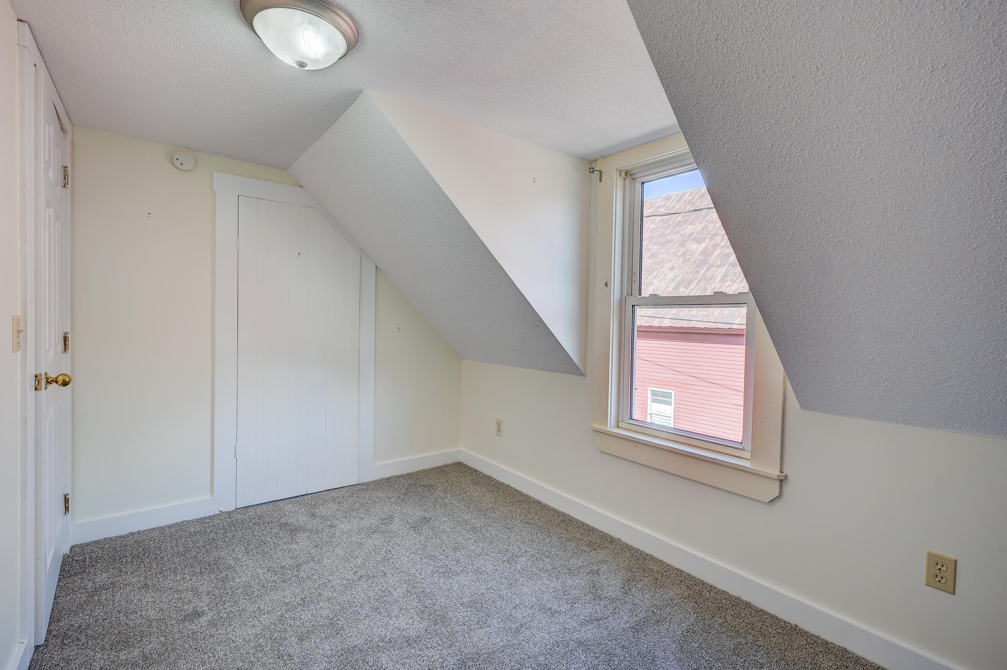 Empty carpeted attic room with a sloped ceiling, closet door, and window.