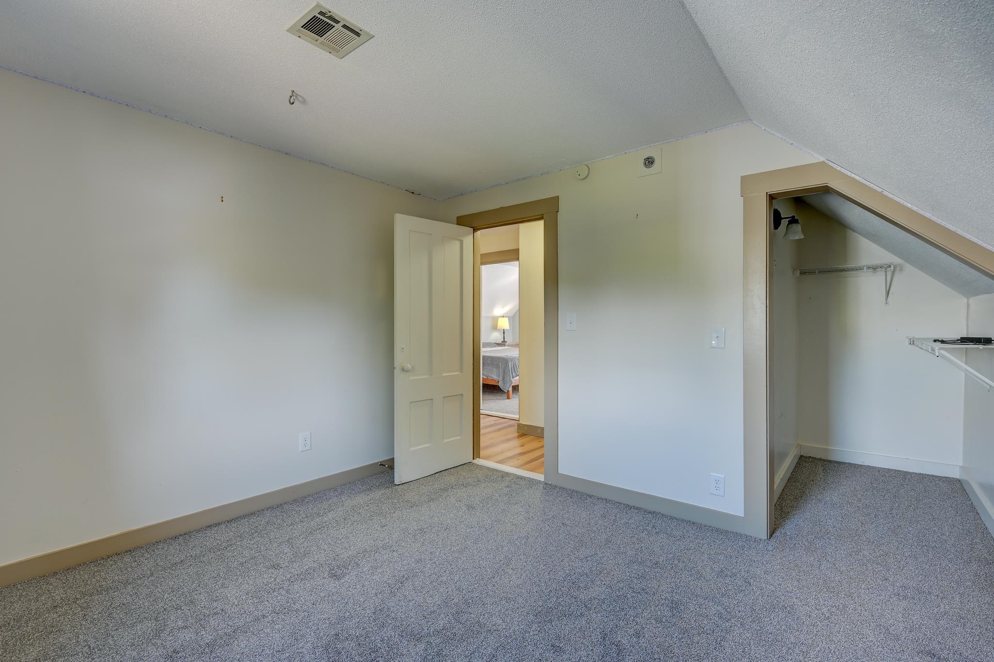 Vacant carpeted room with a sloped ceiling and open closet nook, looking through the doorway into a bedroom.