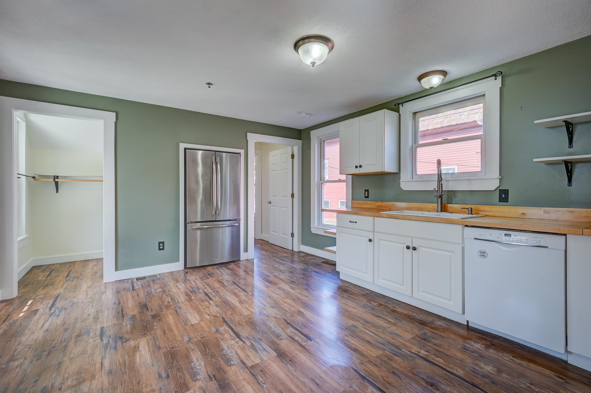 Kitchen with white cabinets, stainless steel refrigerator, sink beneath two windows, and wood-look flooring.