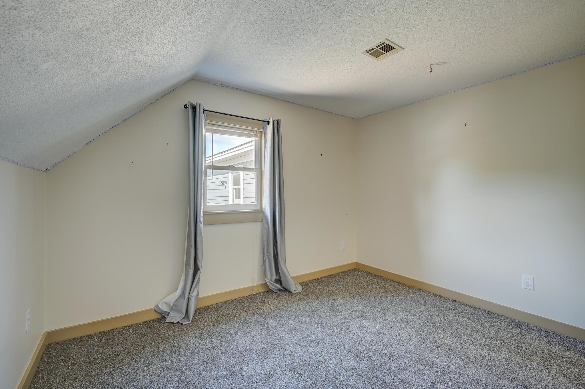 Empty bedroom with gray carpet, sloped ceiling, and a window framed by long gray curtains.