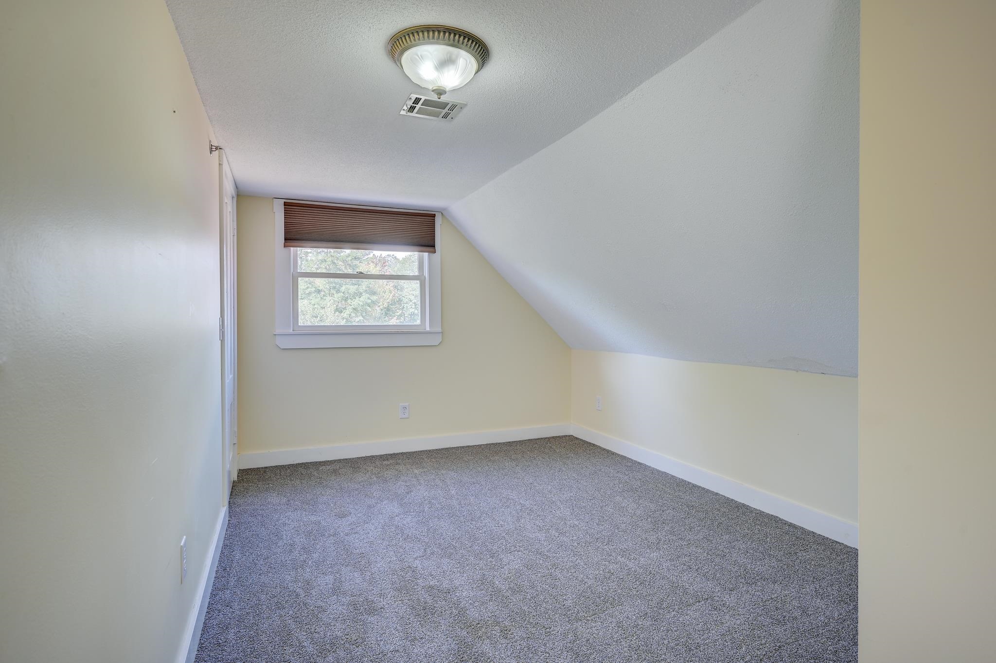 Vacant attic bedroom with gray carpet, a sloped ceiling, and a shaded window.
