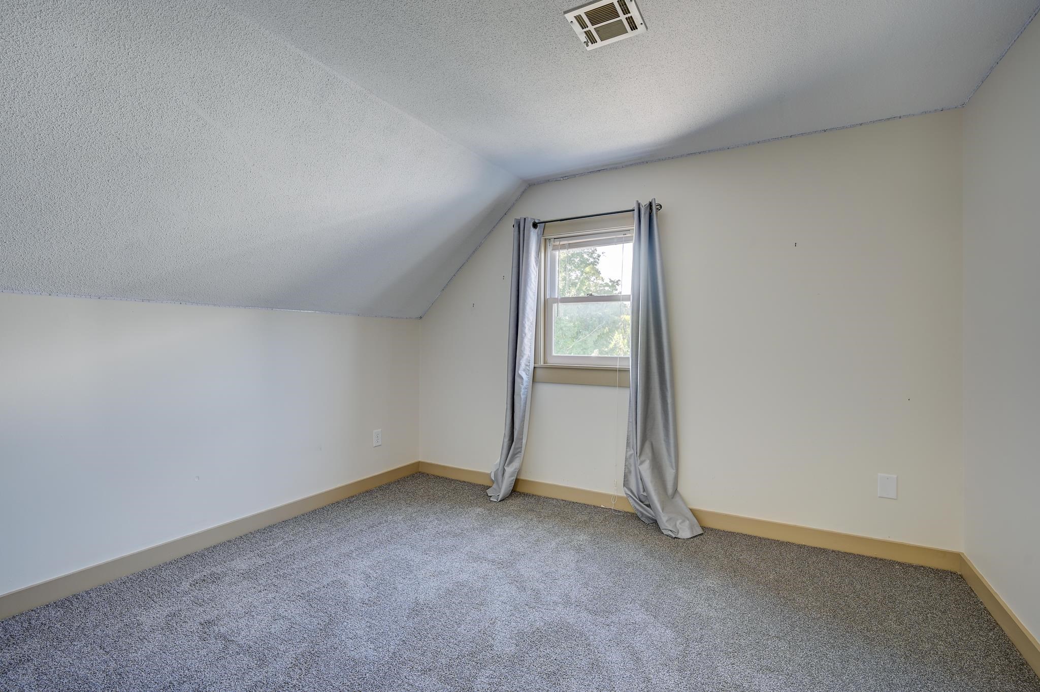Empty bedroom with gray carpet, a sloped ceiling, and a curtained window.