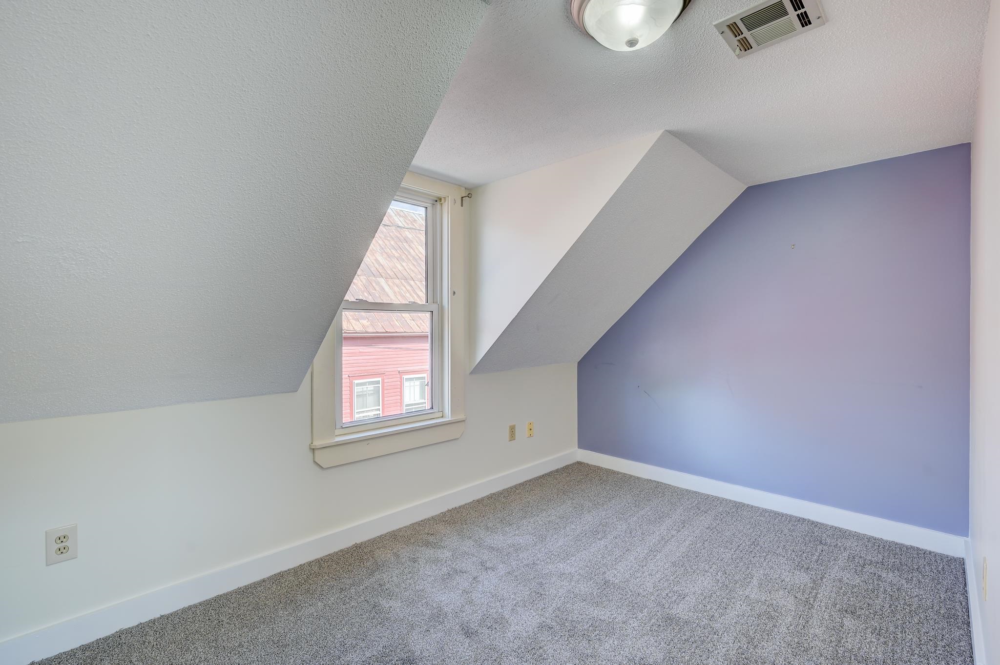 Empty attic bedroom with a lavender wall, gray carpet, sloped ceilings, and a window.