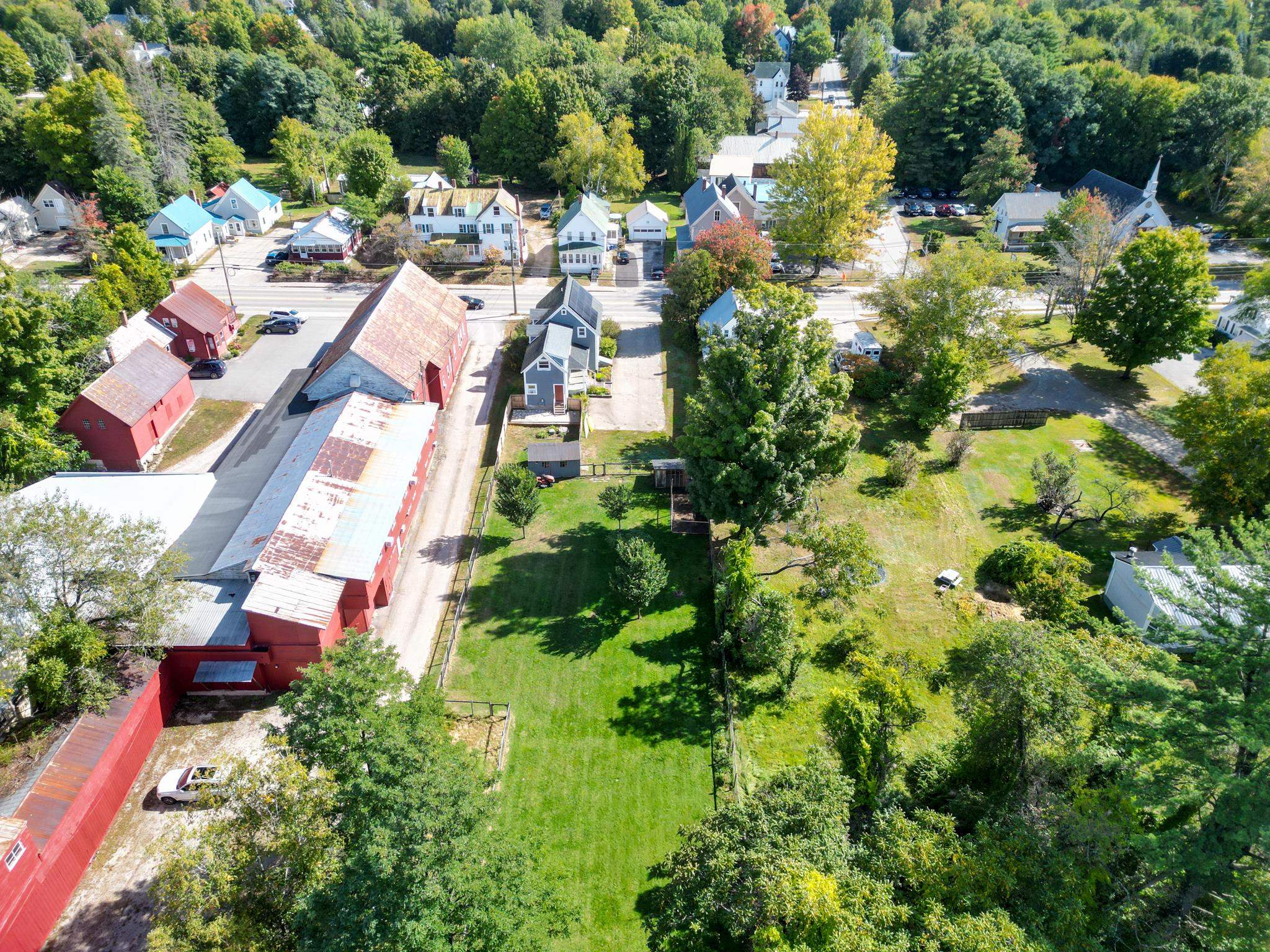 Aerial view of a gray-blue house beside a long lawn, mature trees, and neighboring buildings.