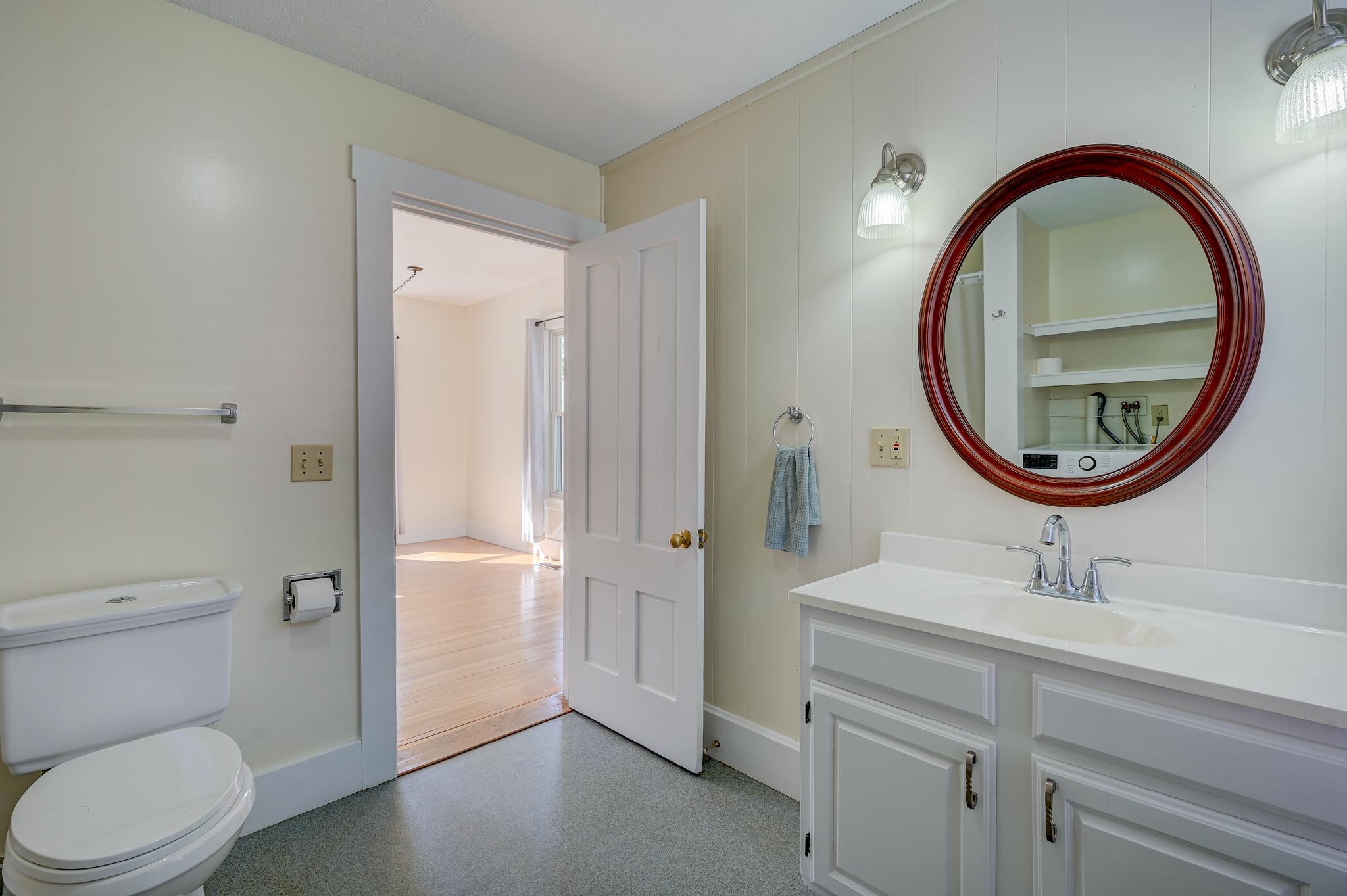 Bathroom with a white vanity, oval wood-framed mirror, and open door to a bright room.