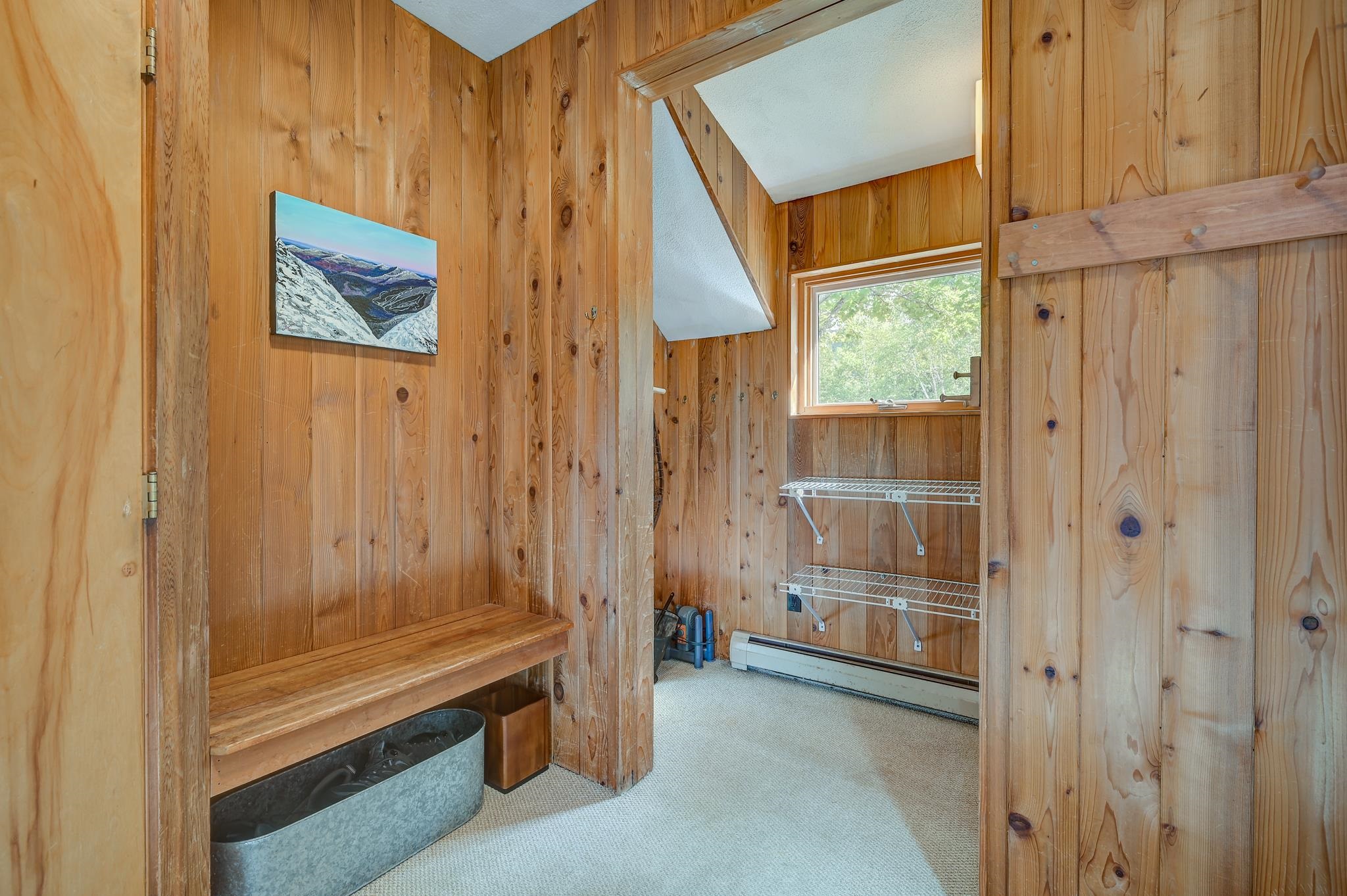 Wood-paneled mudroom with a built-in bench, boot tray, and drying racks beside a window.