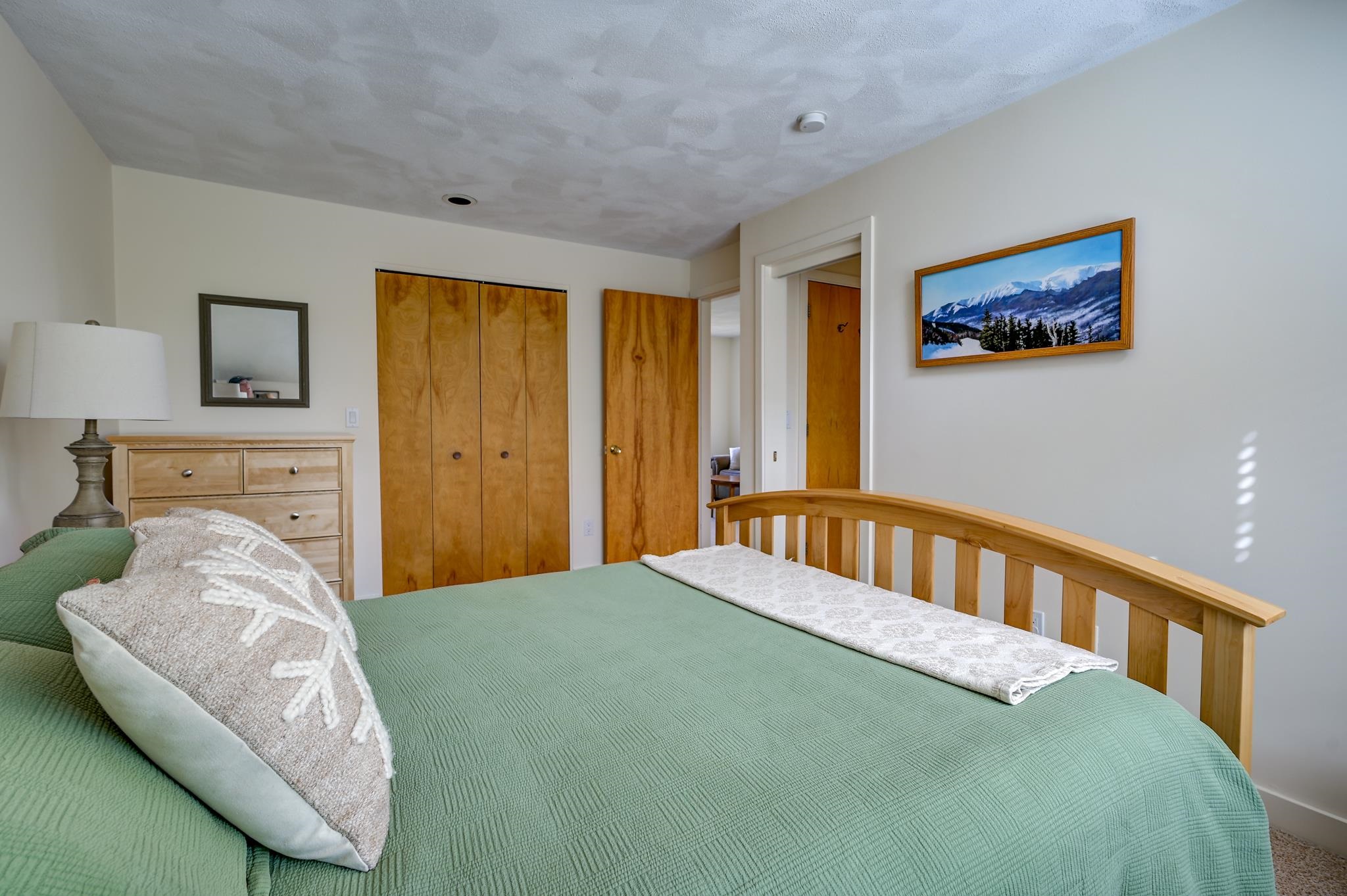 Bedroom with a green quilted bed, wooden headboard, and framed mountain landscape.