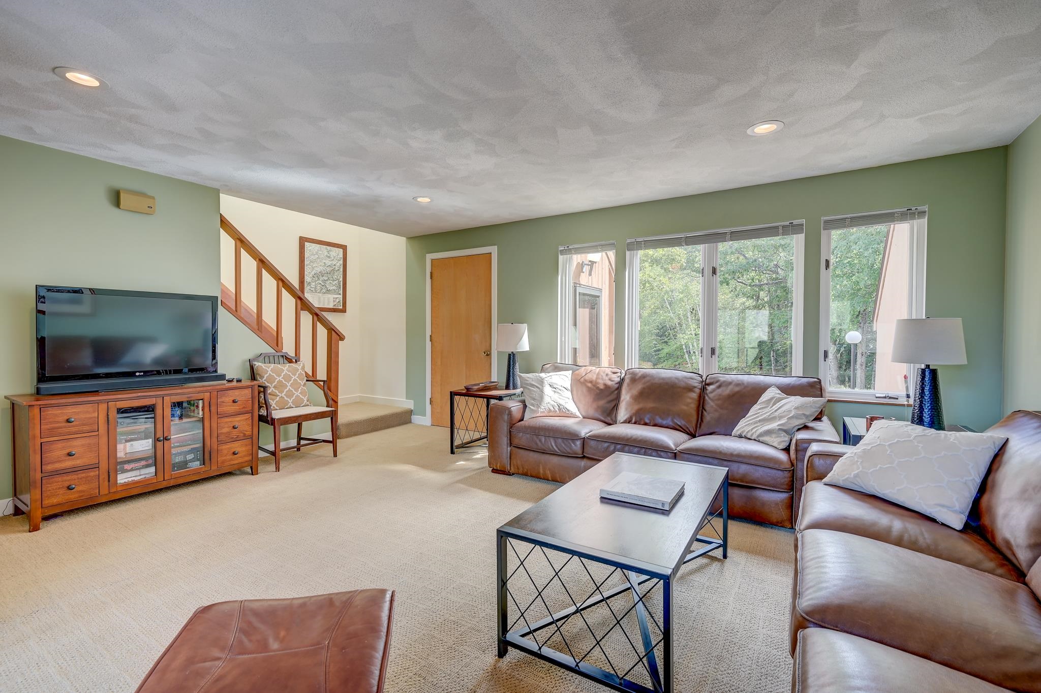 Living room with brown leather sofas, a metal coffee table, and windows overlooking trees.