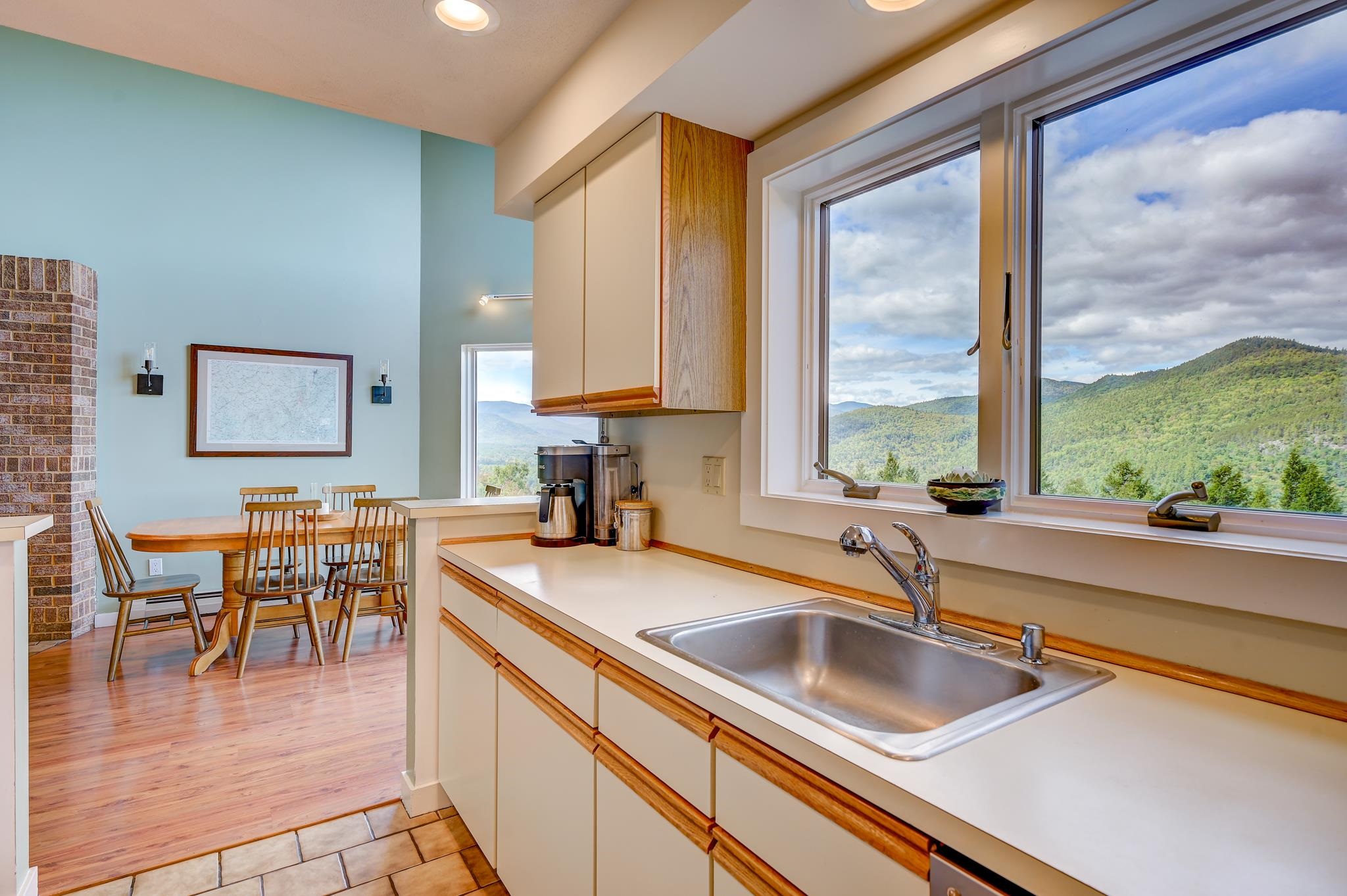 Kitchen sink and wood-trimmed cabinets beside windows overlooking forested mountains.