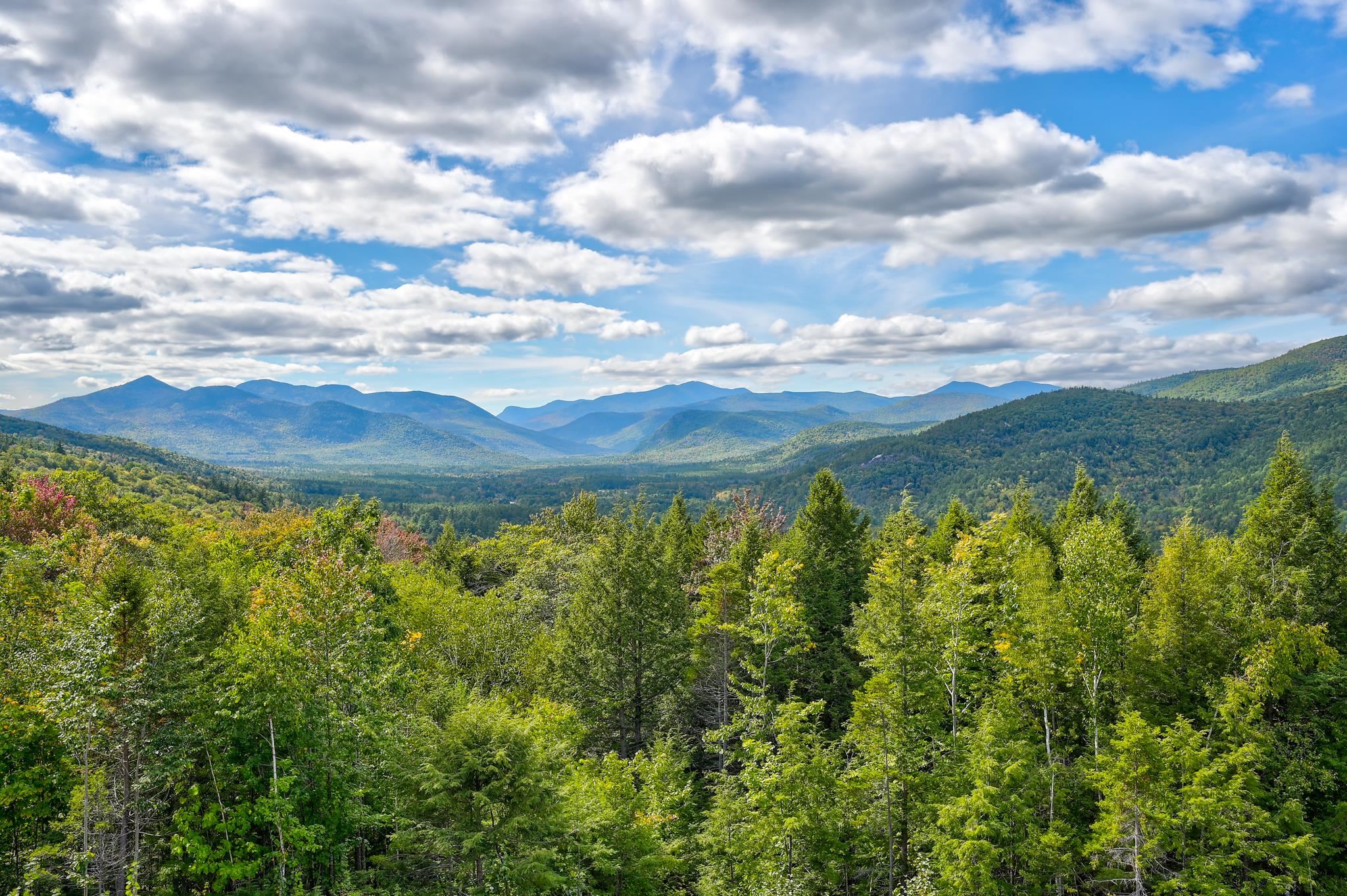 Layered forested mountains and a green valley beneath a cloud-filled blue sky.