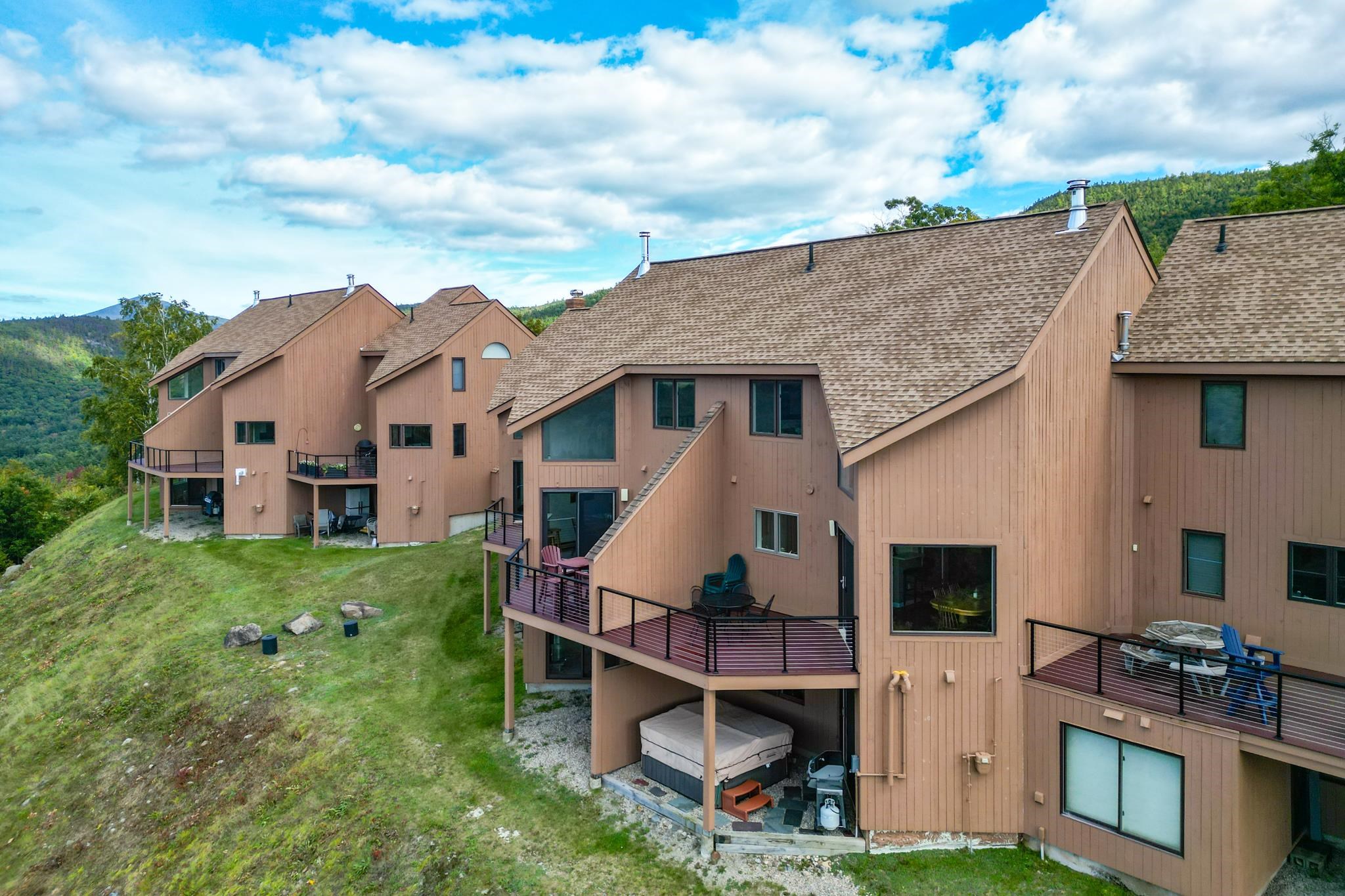 Attached brown mountain condos with private decks on a grassy hillside beneath wooded hills.