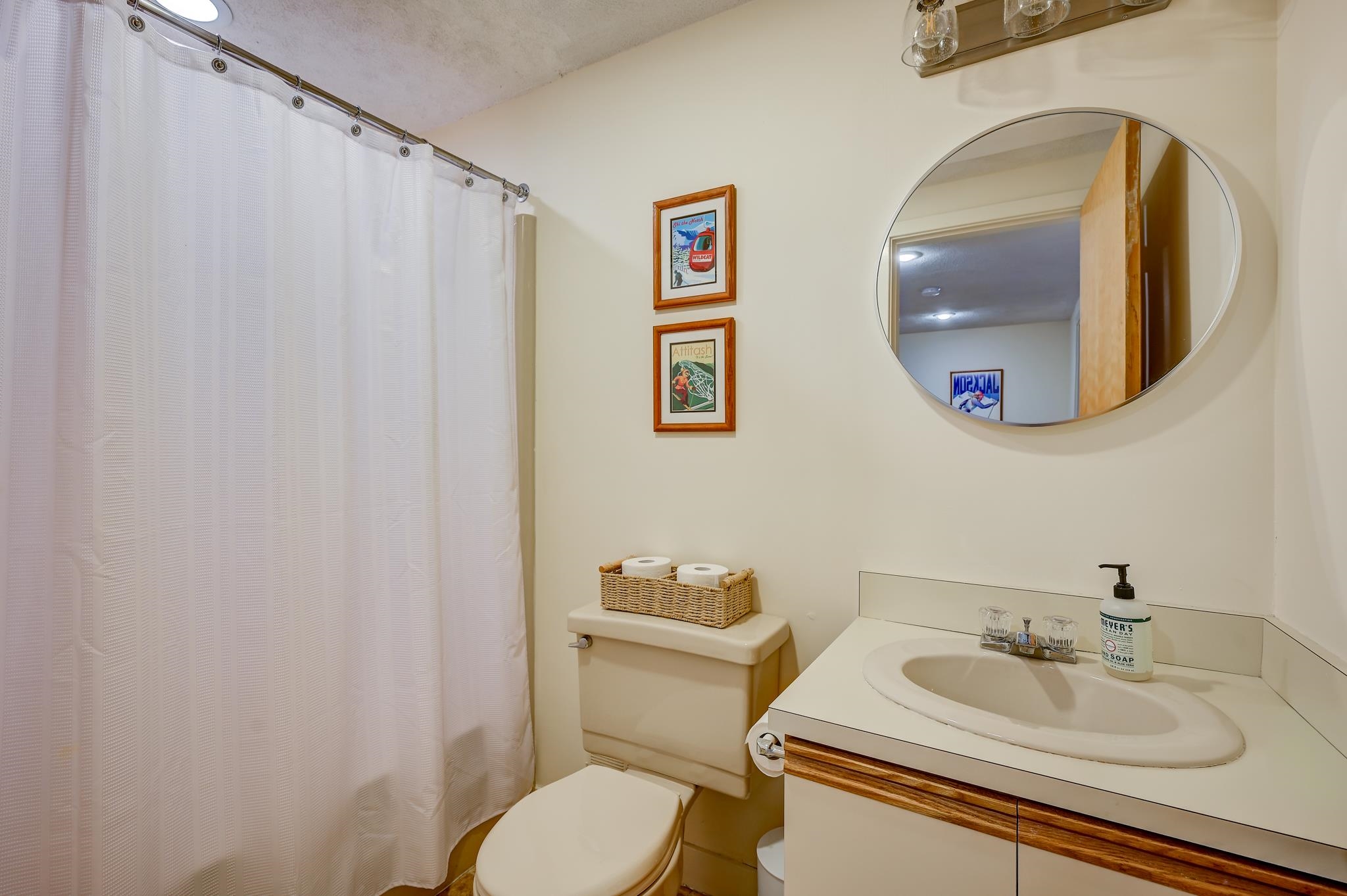 Bathroom with a white shower curtain, wicker basket on the toilet, and cream vanity beneath a round mirror.