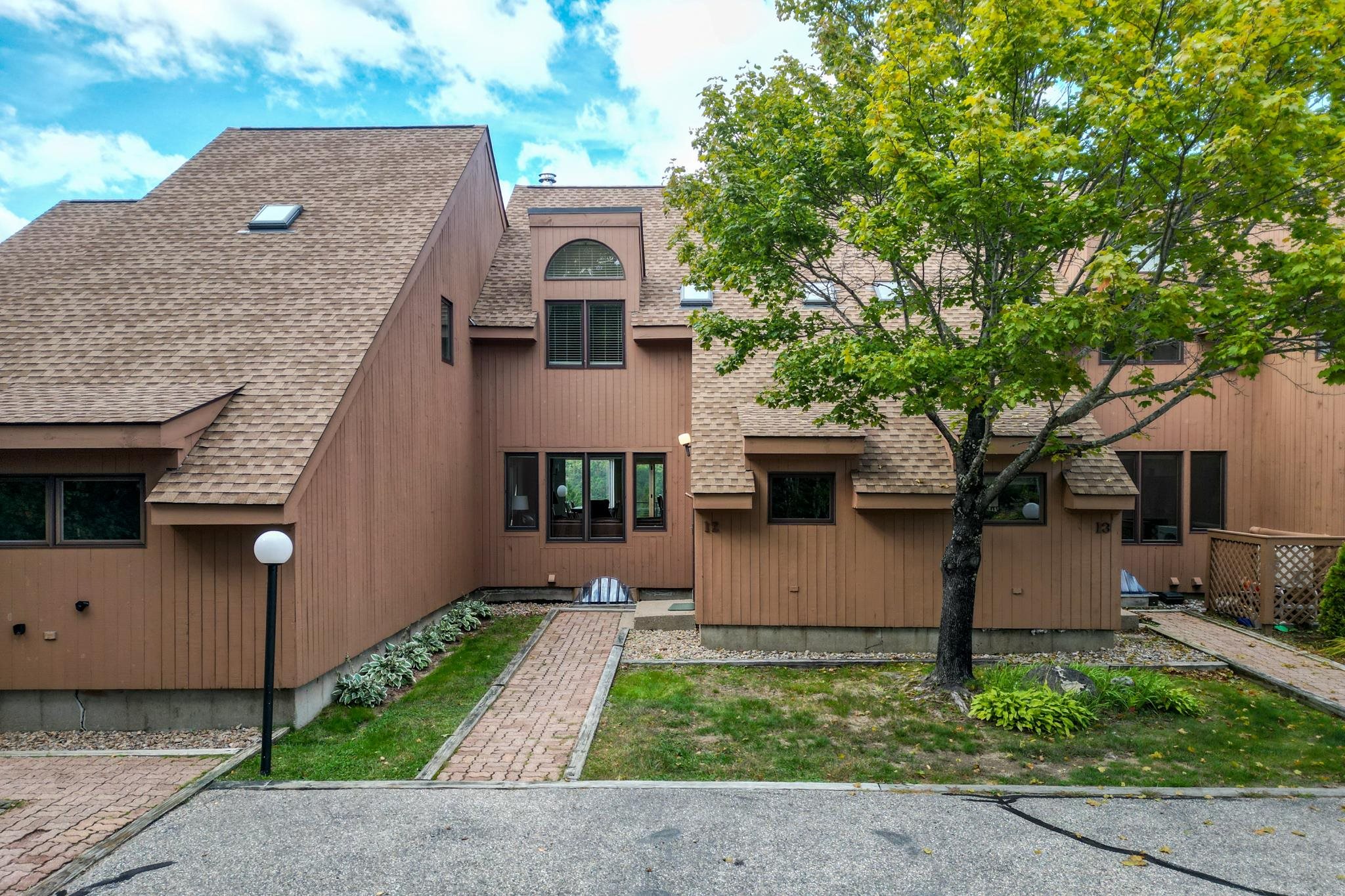 Brown-sided condominium exterior with steep shingled roofs, a brick walkway, and a mature tree.