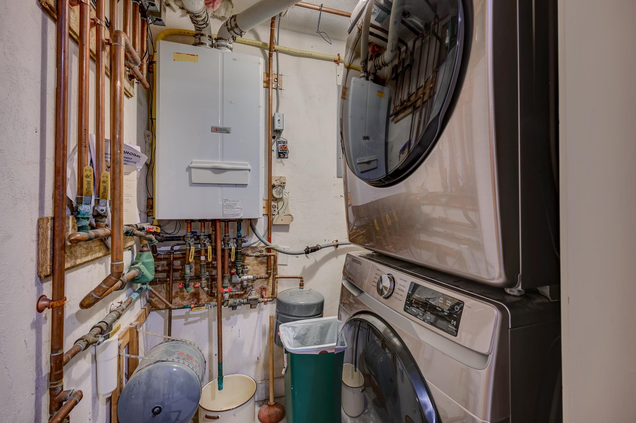 Utility room with stacked washer and dryer beside heating equipment and exposed copper pipes.