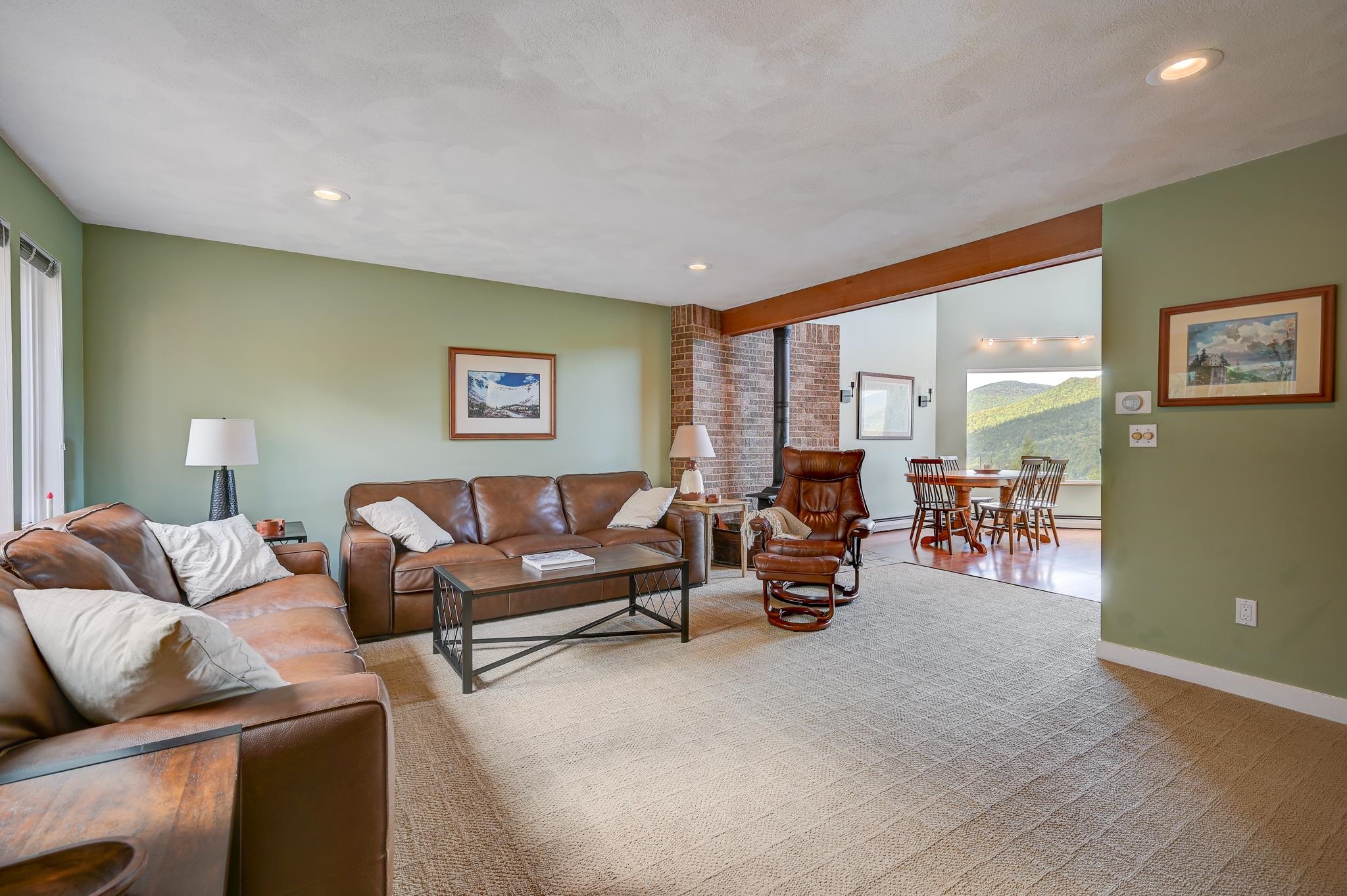 Living room with brown leather seating, a brick fireplace, and a dining area overlooking wooded mountains.