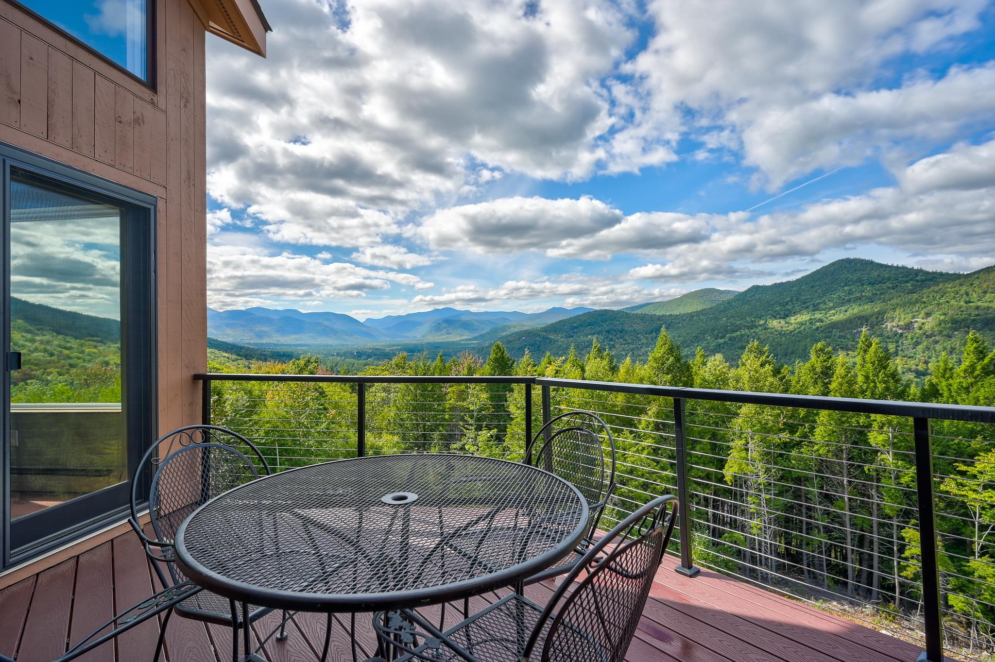 Furnished deck overlooking a forested mountain valley, with a round metal table and cable railing.