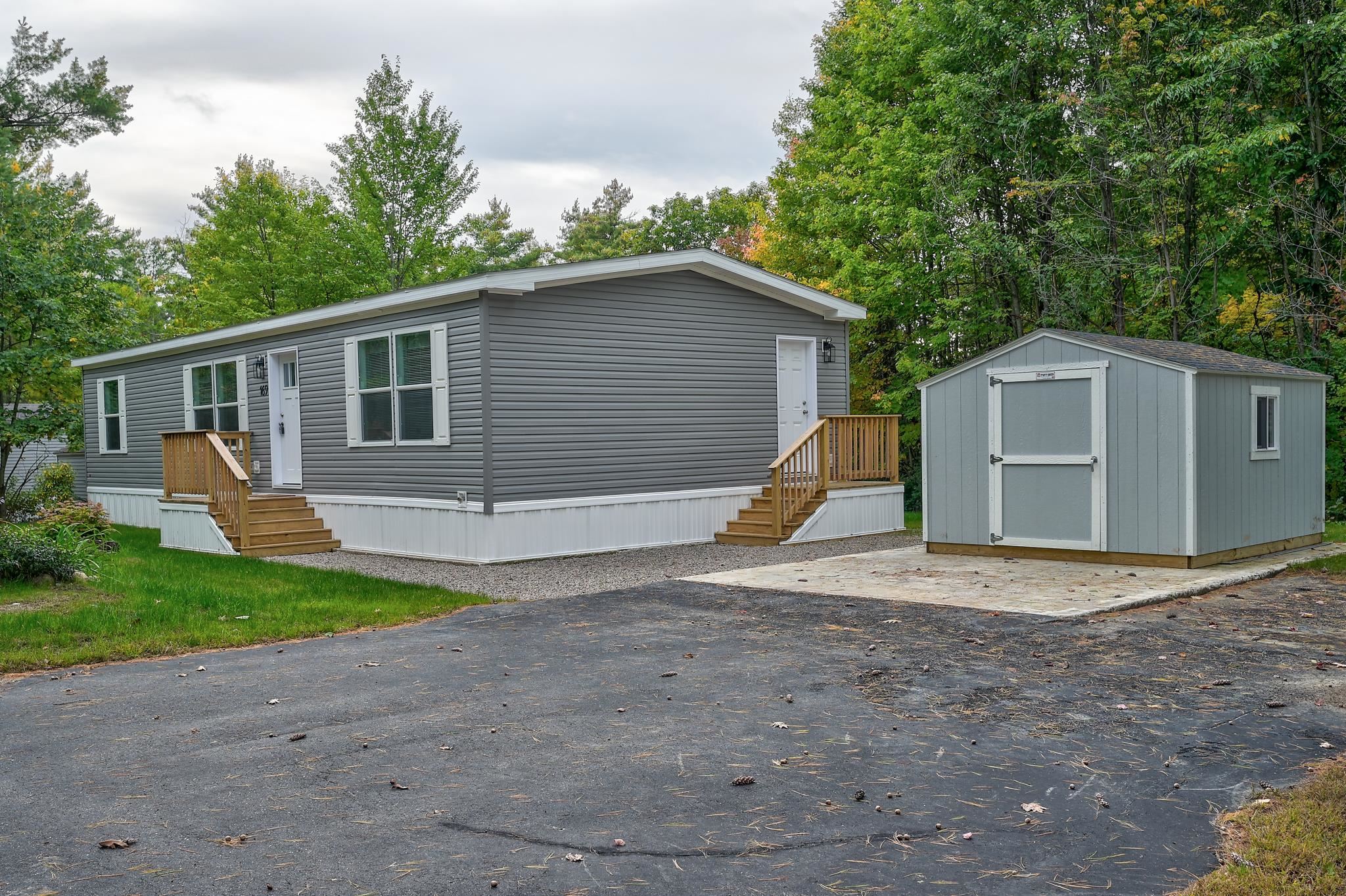 Gray manufactured home with two wooden stairways and a matching storage shed beside the driveway.