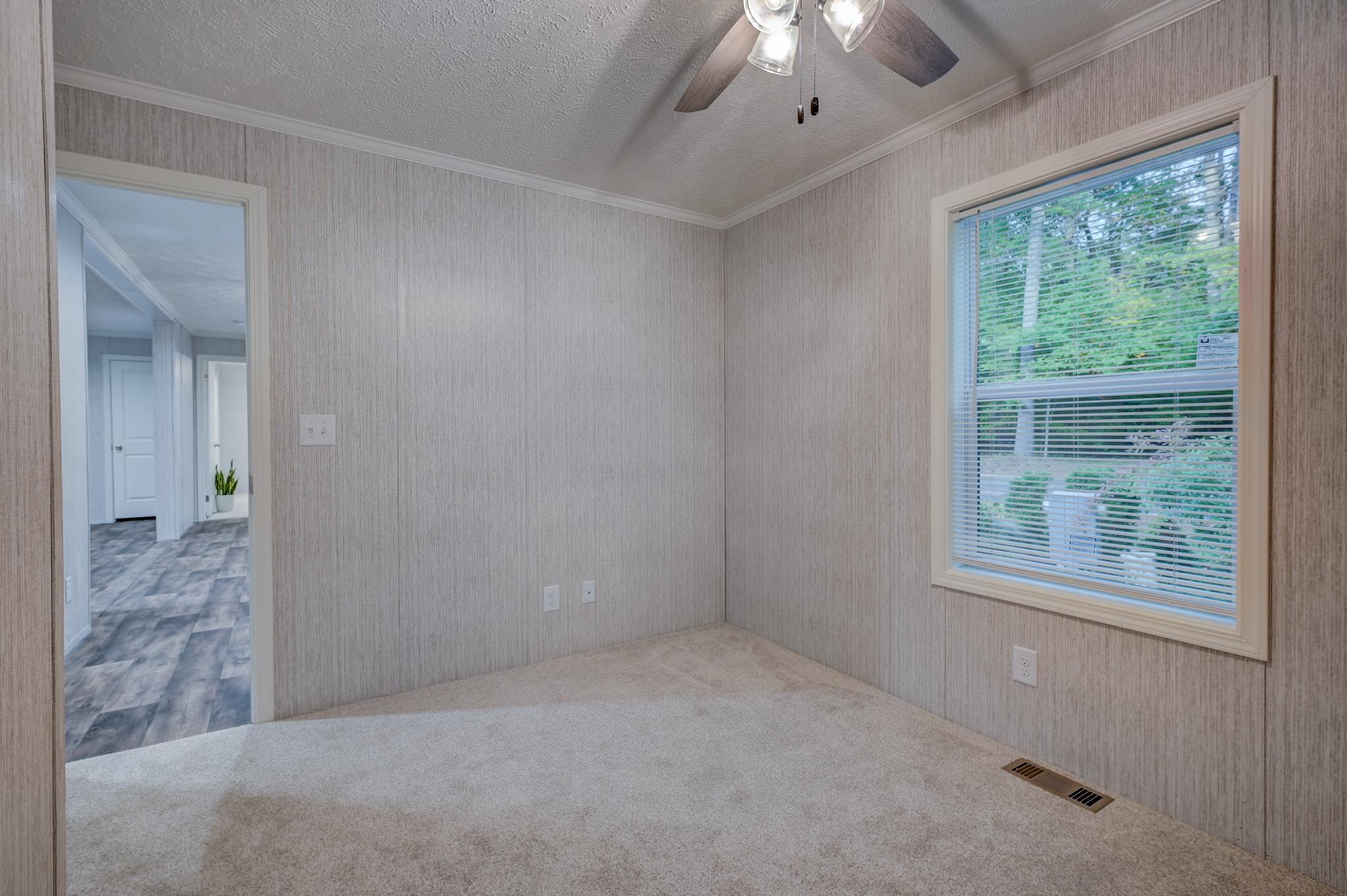 Empty carpeted room with a ceiling fan and a large window overlooking greenery.