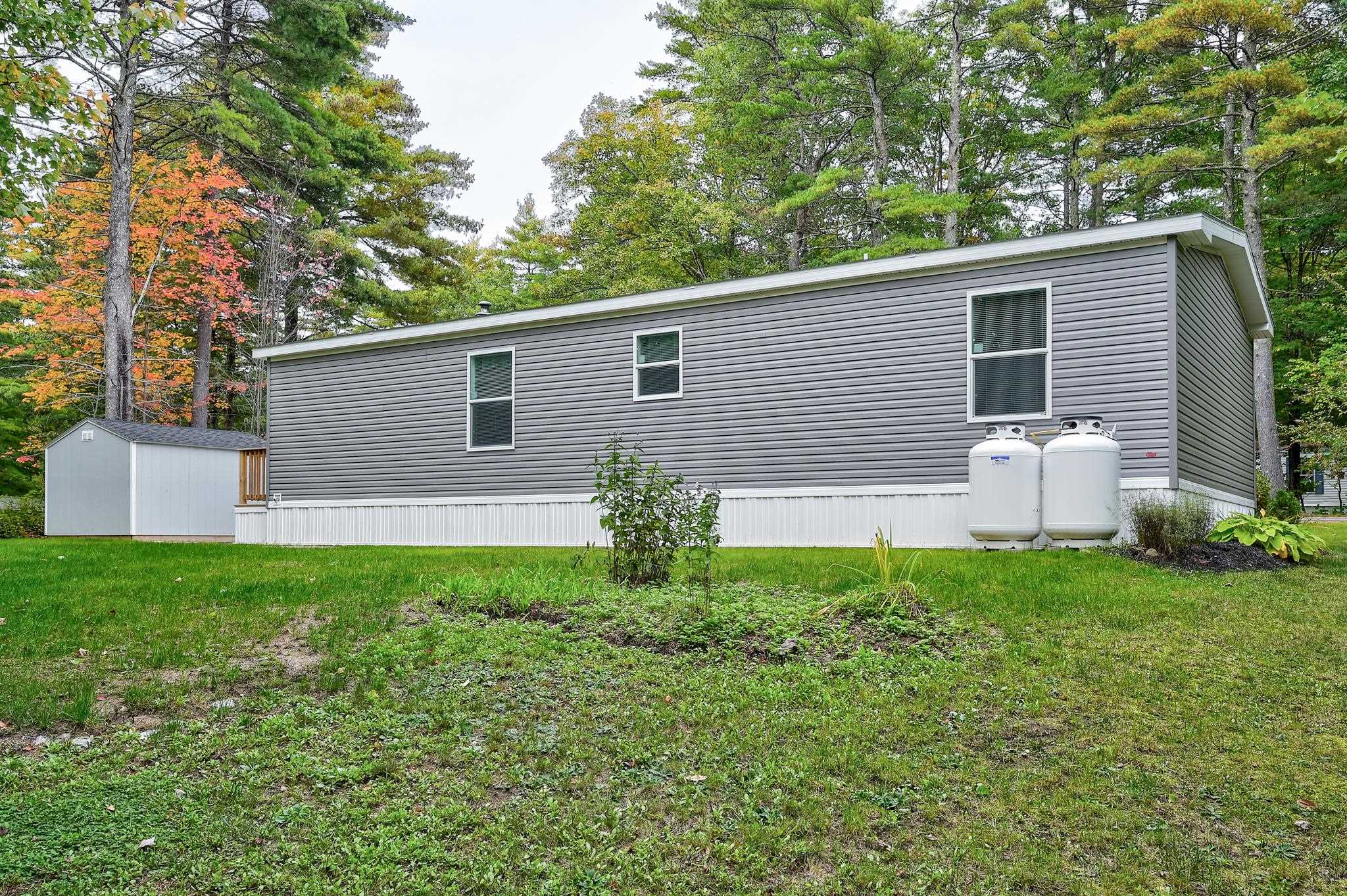 Gray manufactured home with two propane tanks and a small shed on a wooded lot.