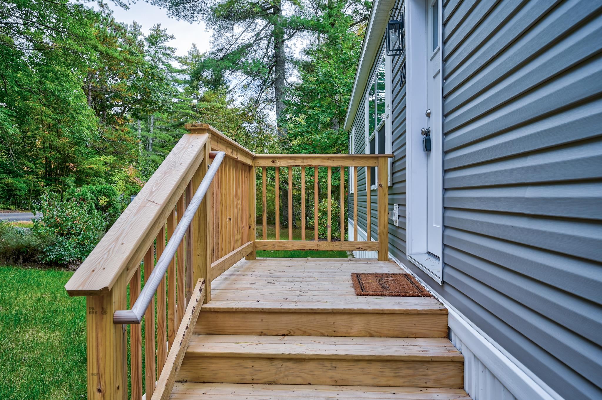 Wooden entrance steps and landing beside gray siding, with a leafy wooded yard beyond.