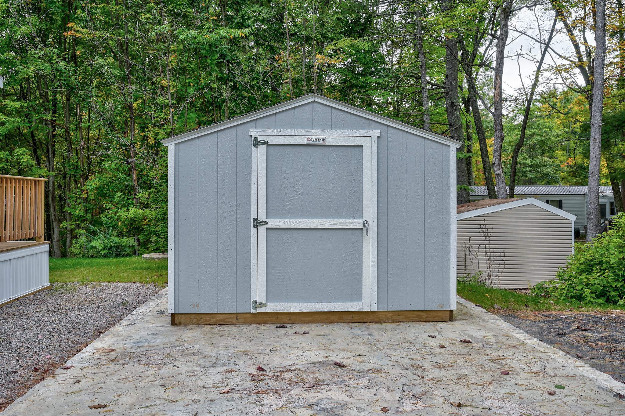 Gray storage shed with white trim and a centered door on a concrete pad in a wooded yard.