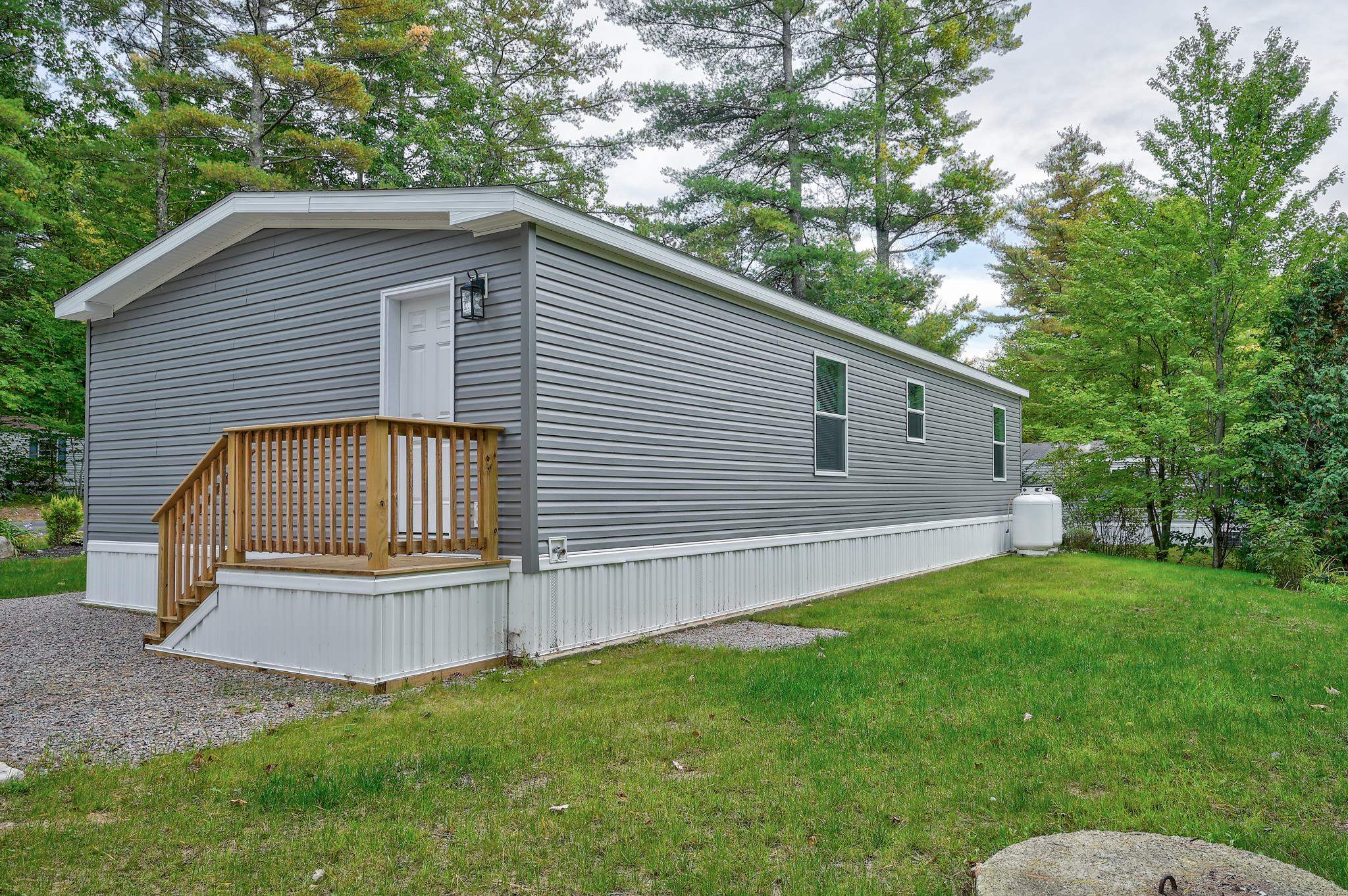Gray manufactured home with a small wooden porch, surrounded by grass and mature trees.