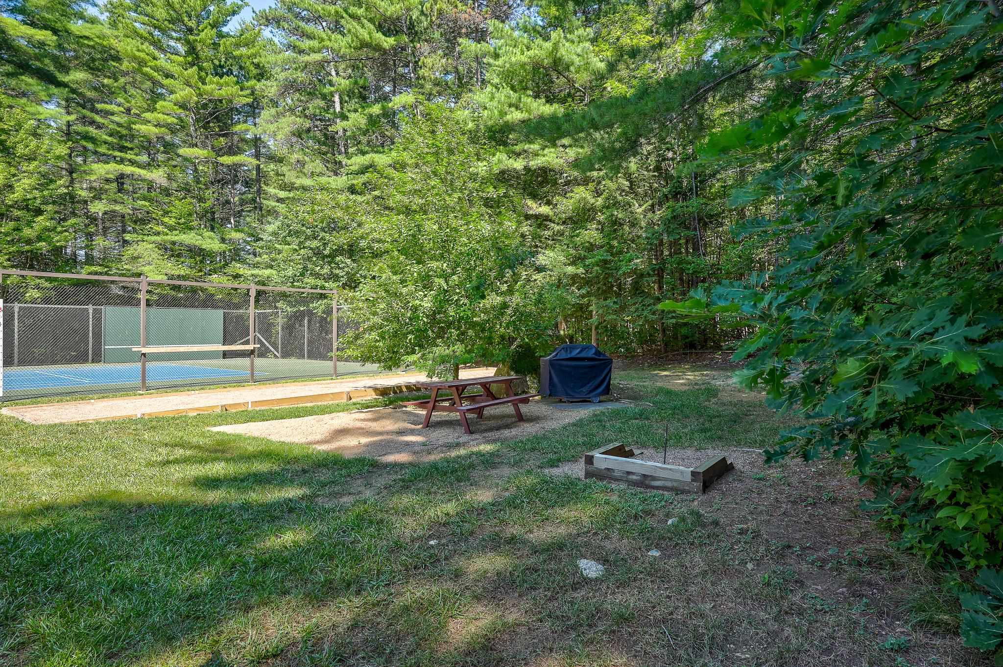 Fenced tennis court beside a wooded picnic area with a table, covered grill, and fire pit.