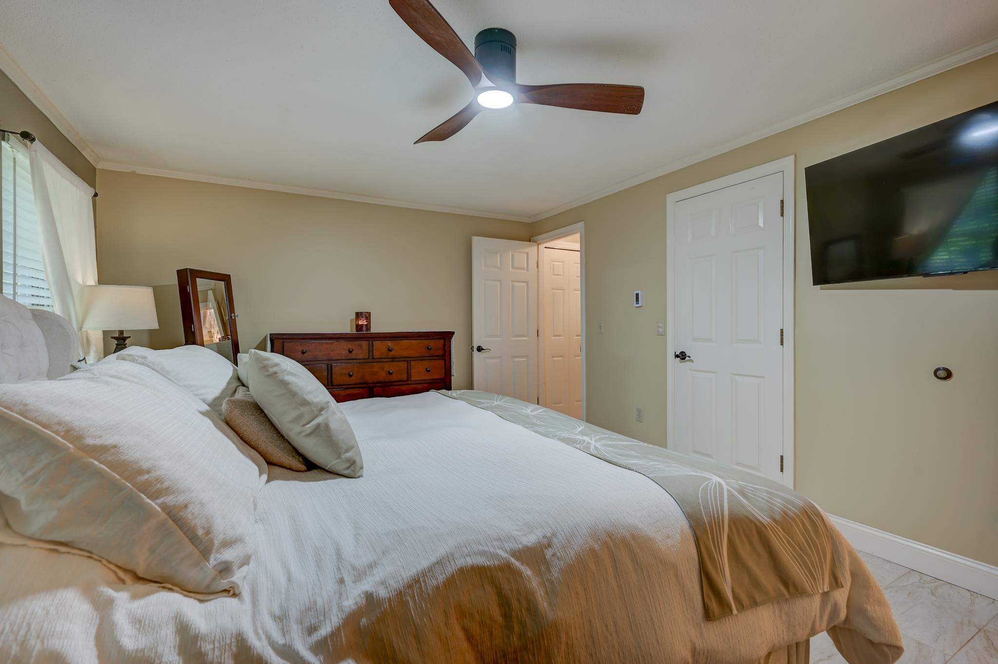 Furnished bedroom with a white bed, dark wood dresser, ceiling fan, and wall-mounted television.