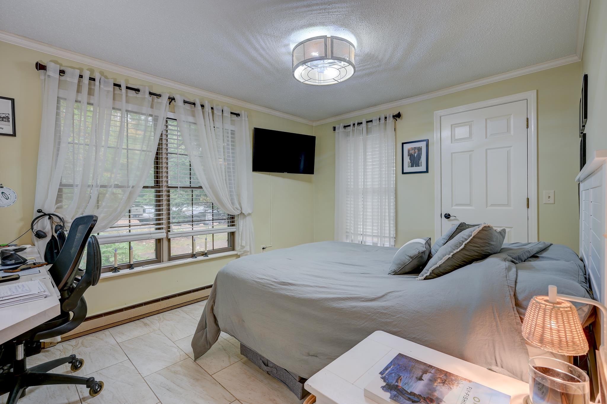 Bedroom with a gray bed, desk and chair, wall-mounted TV, and wide curtained windows.