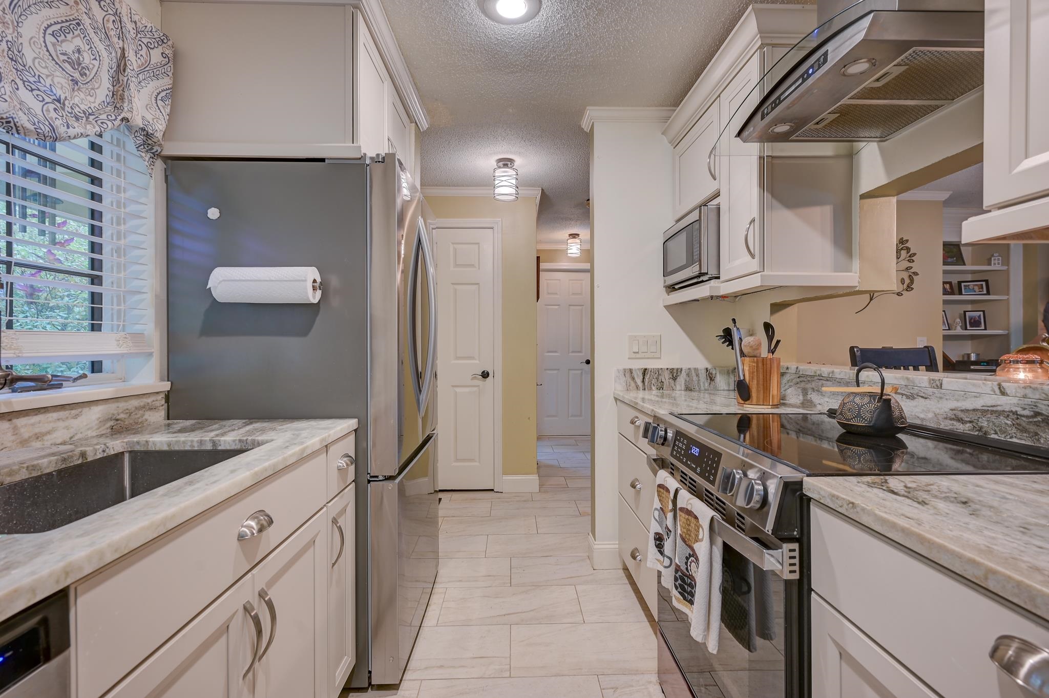 Galley kitchen with white cabinets, marble-pattern counters, stainless appliances, and a window above the sink.