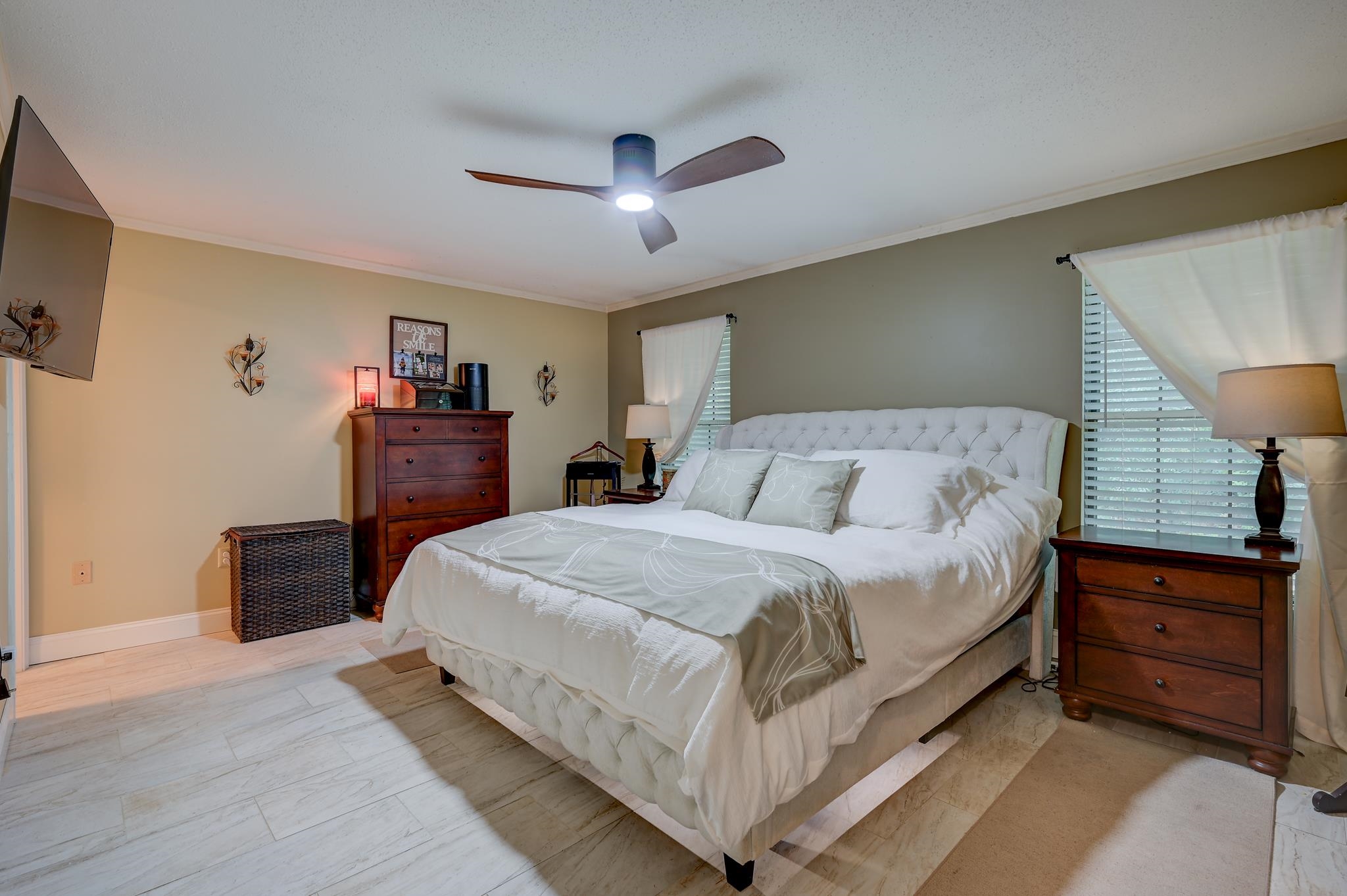 Bedroom with a large white tufted bed, dark wood dressers, and pale tile flooring.