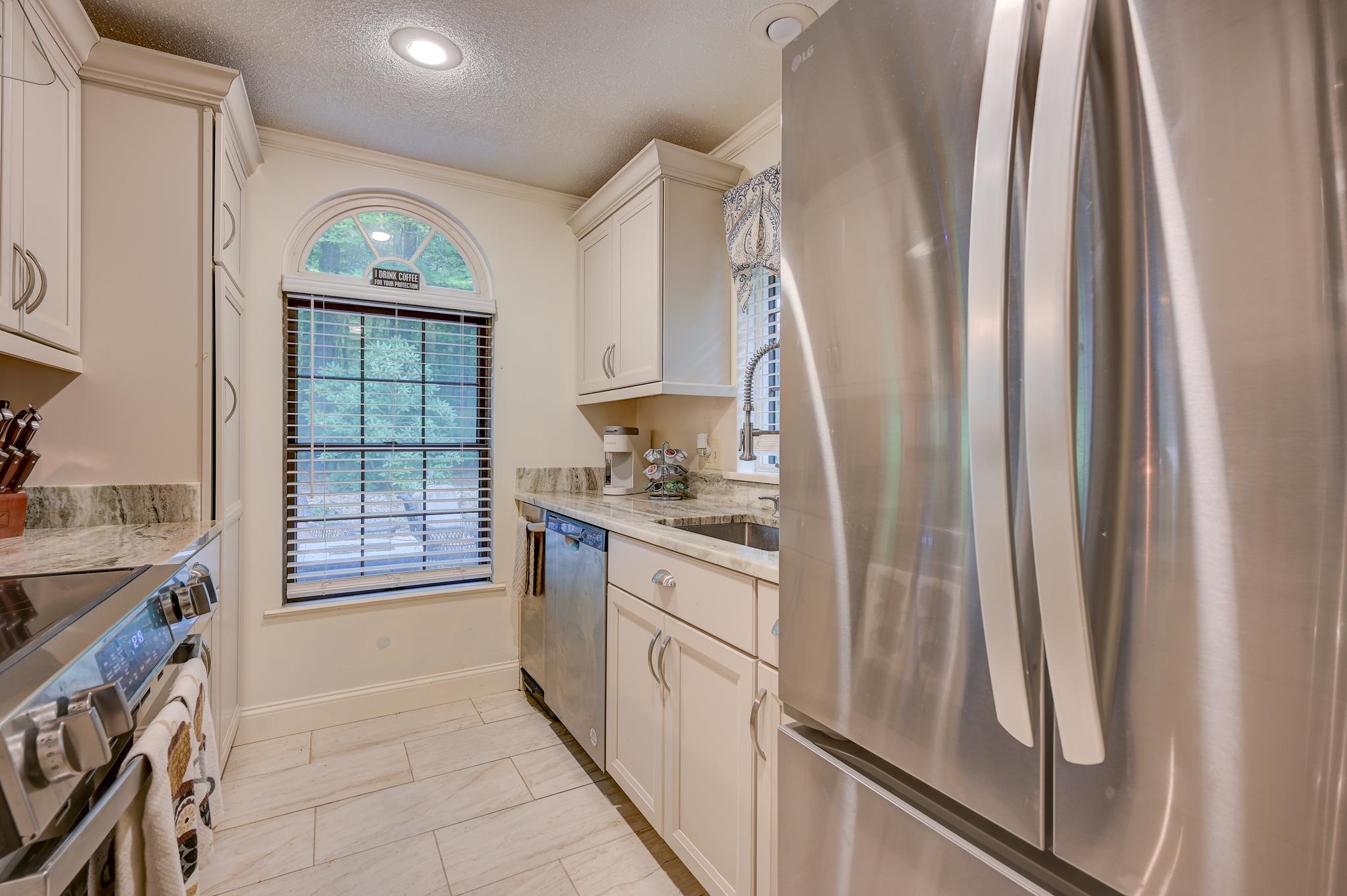 Kitchen with cream cabinets, stone countertops, stainless-steel appliances, and an arched window overlooking trees.