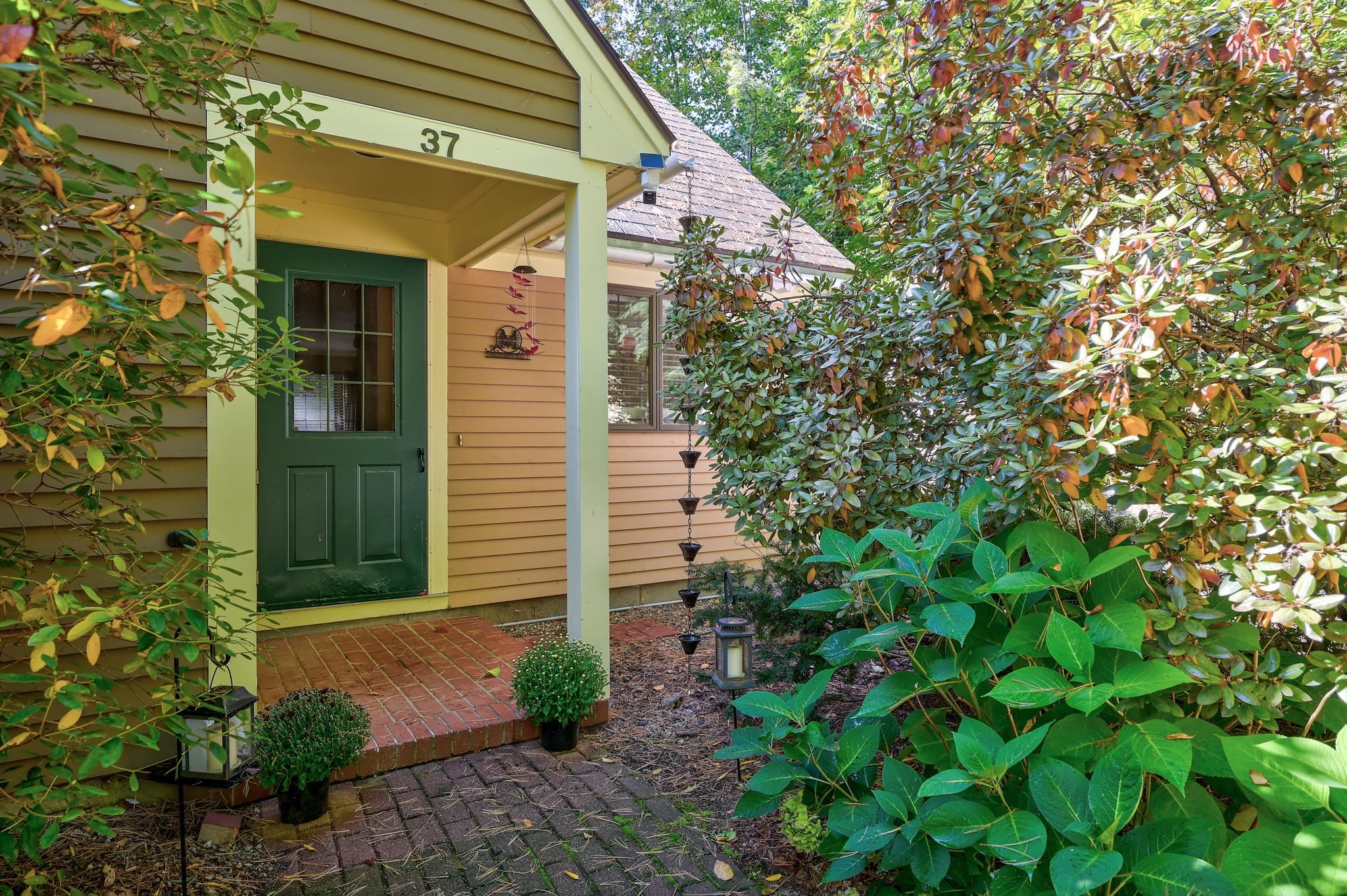 Unit 37 entrance with a dark green door, covered porch, and brick walkway framed by leafy shrubs.