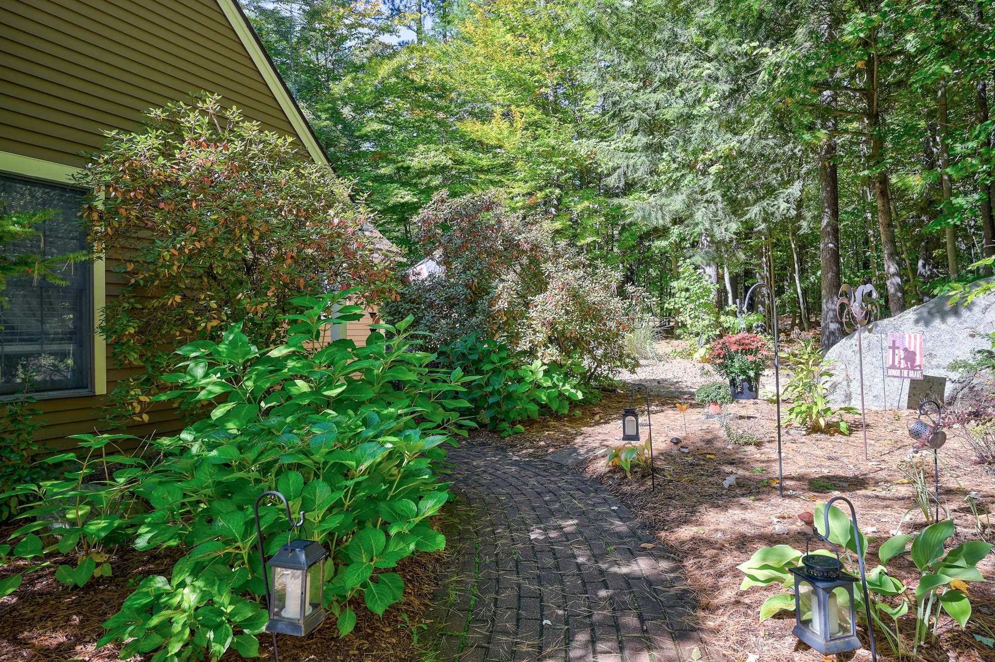 Brick walkway winds through lush garden beds beside a brown-sided home.