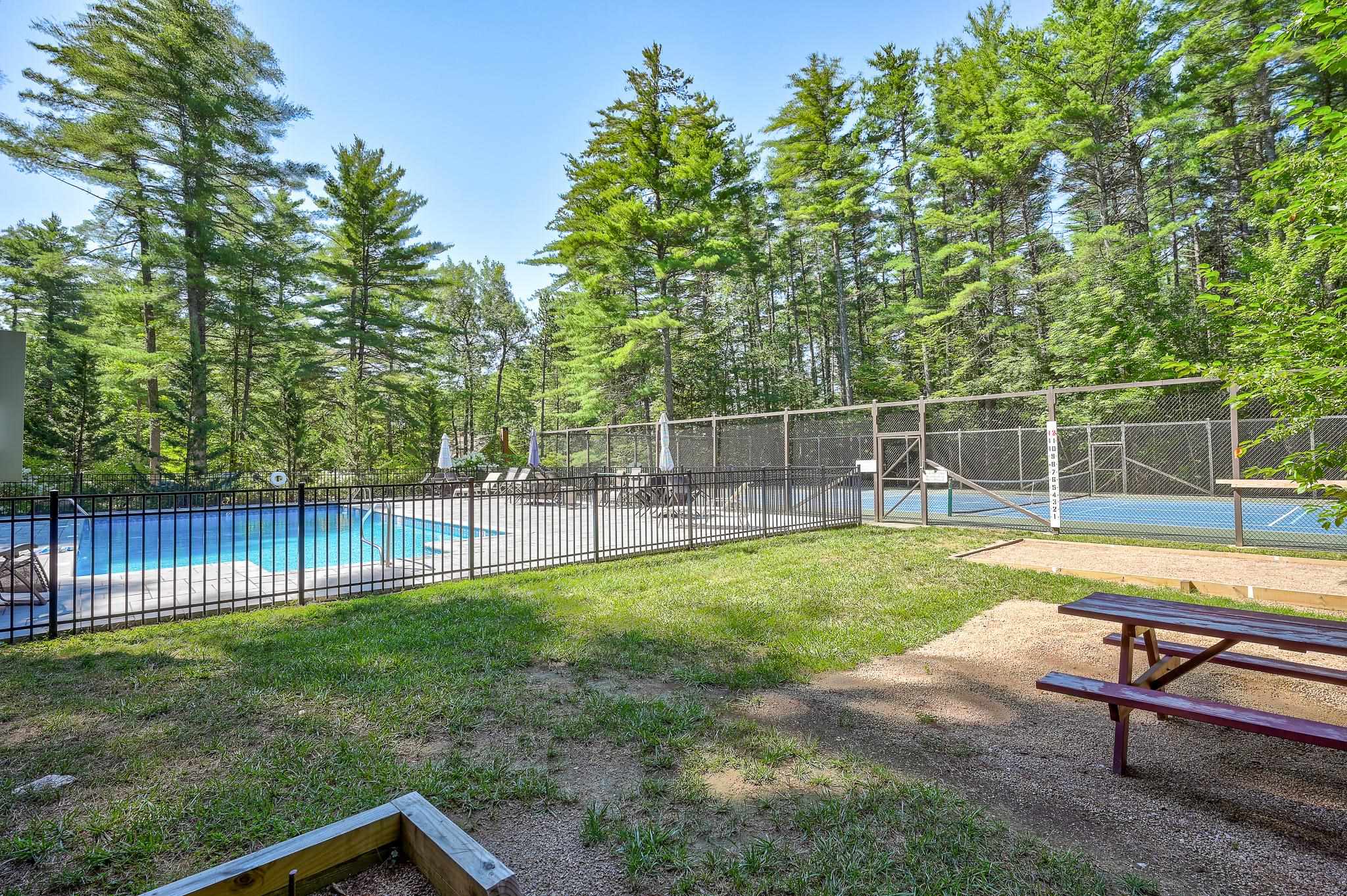 Fenced outdoor pool and tennis court bordered by tall pine trees, with a picnic table nearby.