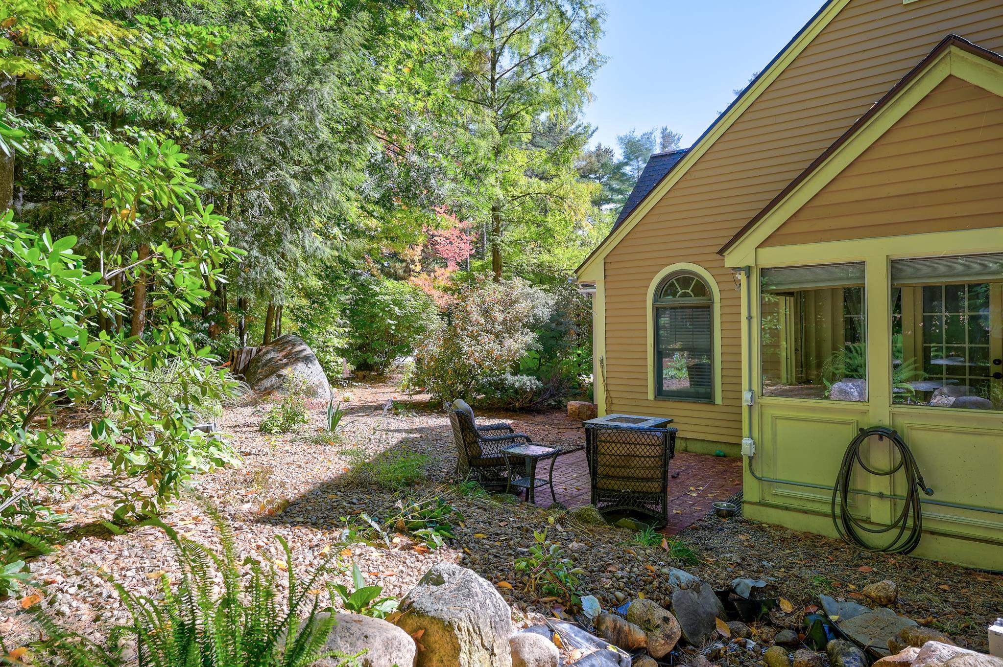 Yellow-sided home beside a brick patio with outdoor seating, surrounded by trees and a rock garden.