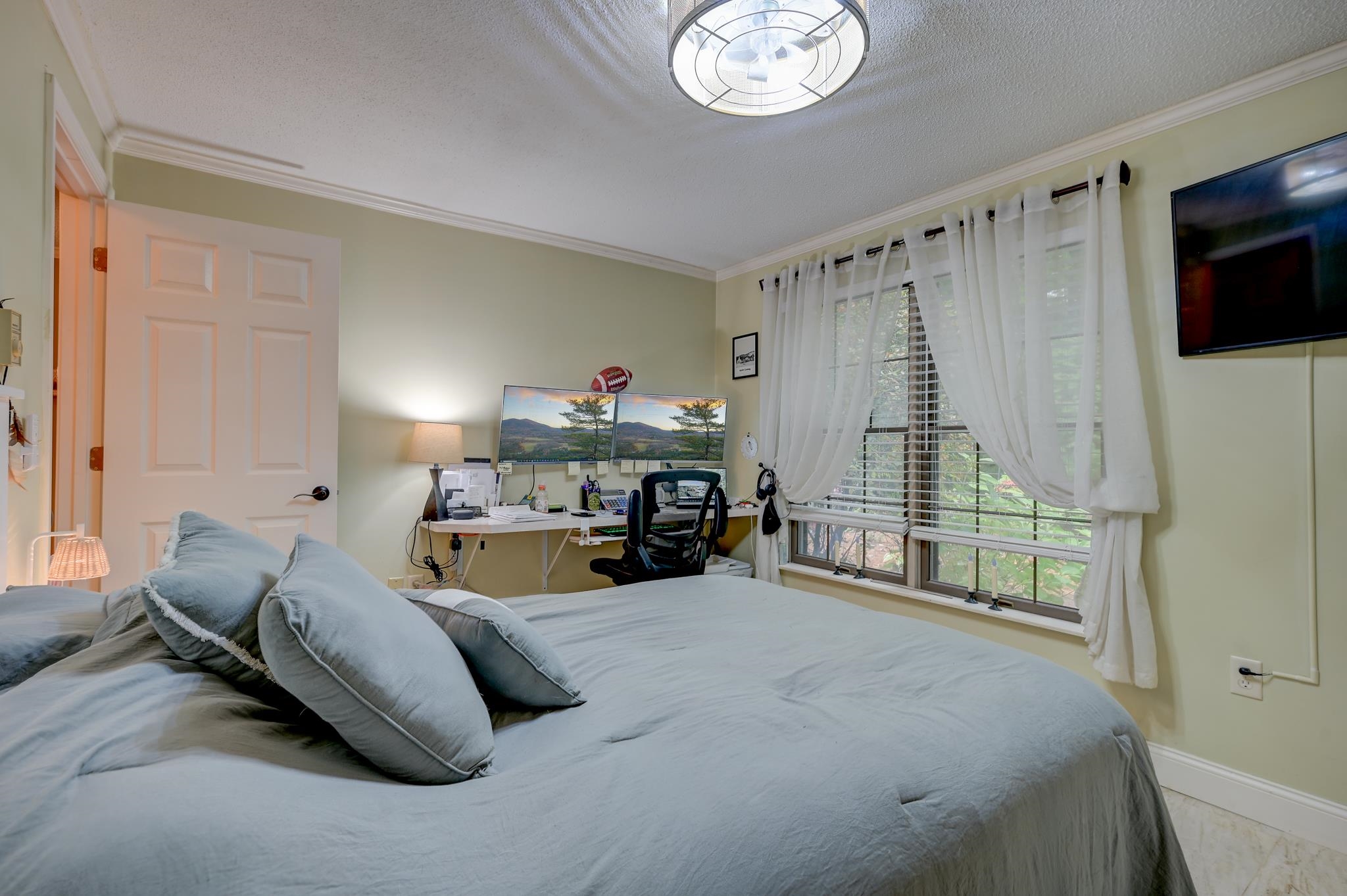 Furnished bedroom with a desk beside a wide window framed by white curtains.