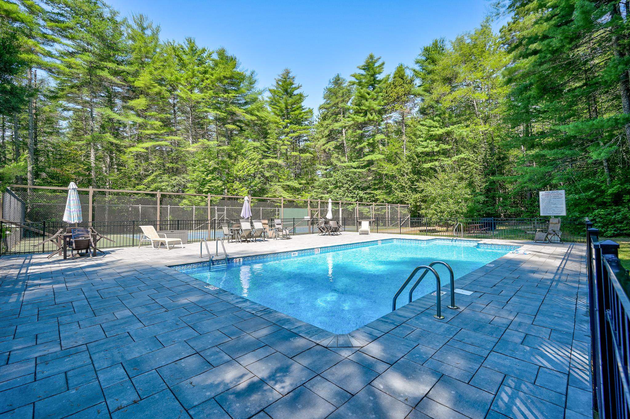 Outdoor pool with a paved deck, lounge chairs, and a wooded backdrop.