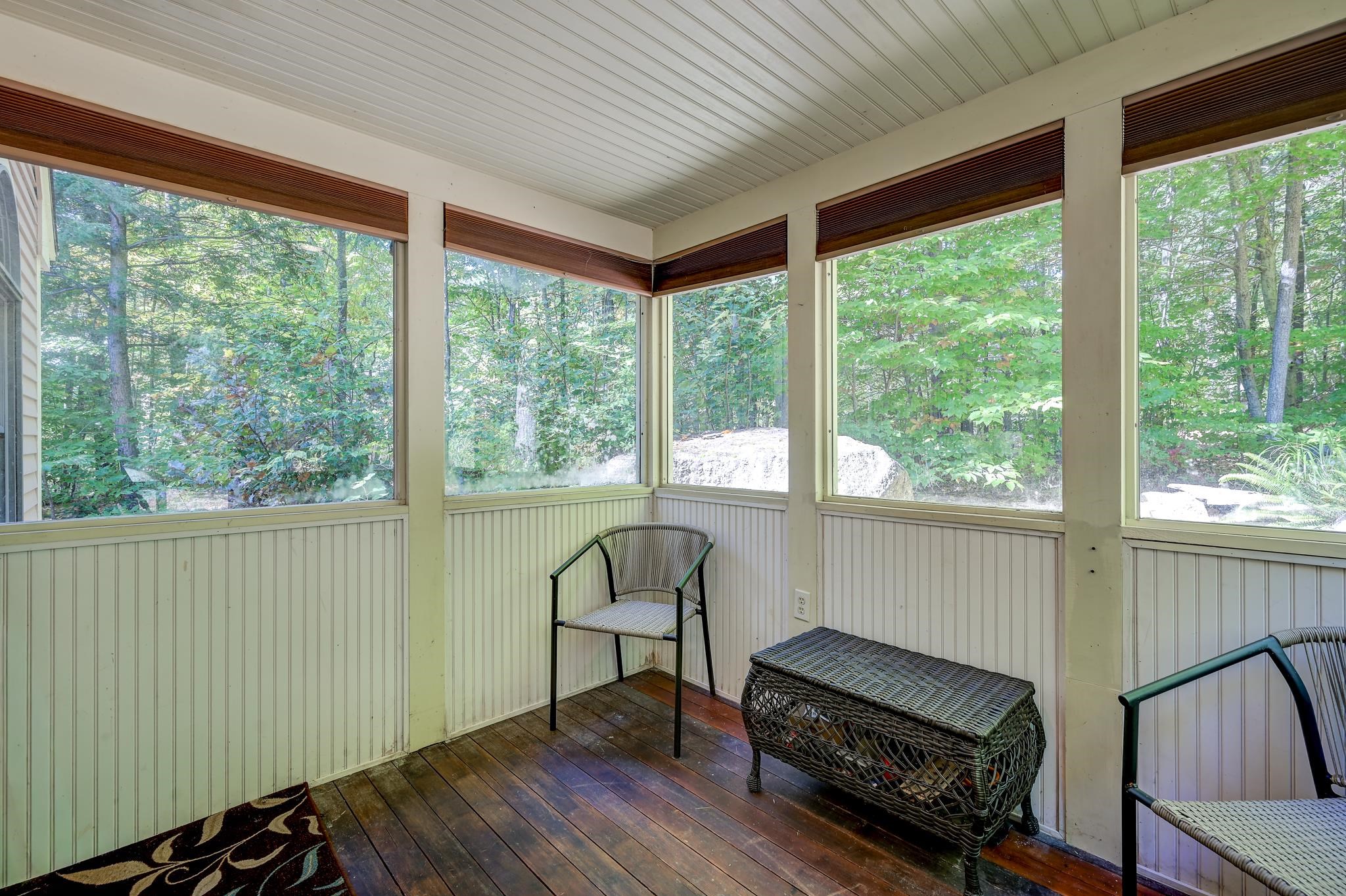 Enclosed porch with wicker seating, dark wood floors, and large windows overlooking leafy woods.