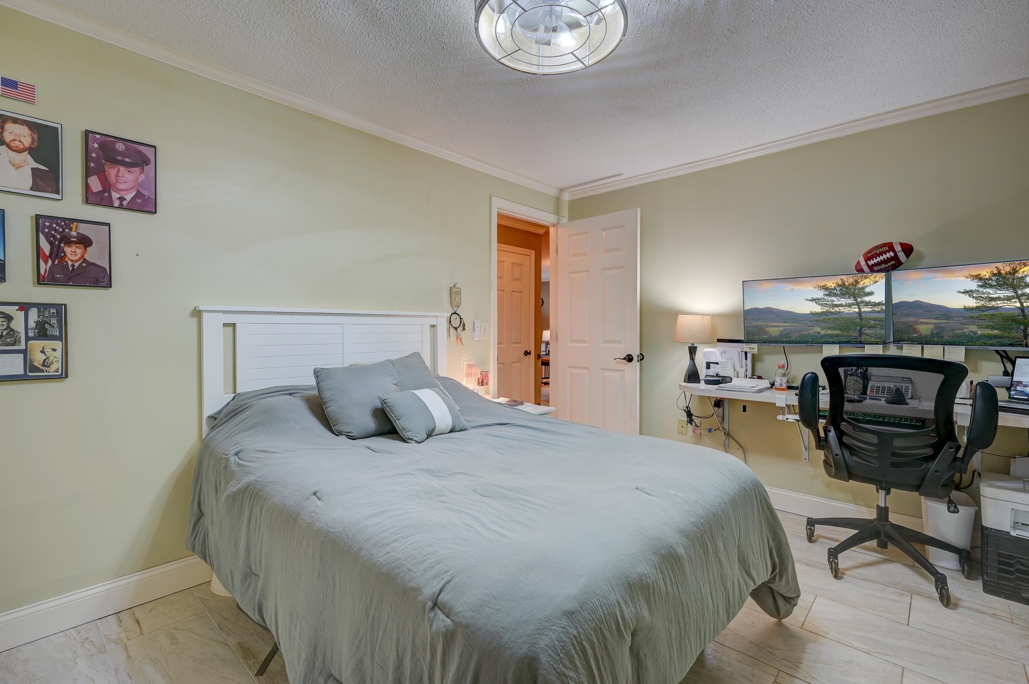Bedroom with a large gray-covered bed and a desk with two monitors showing mountain views.