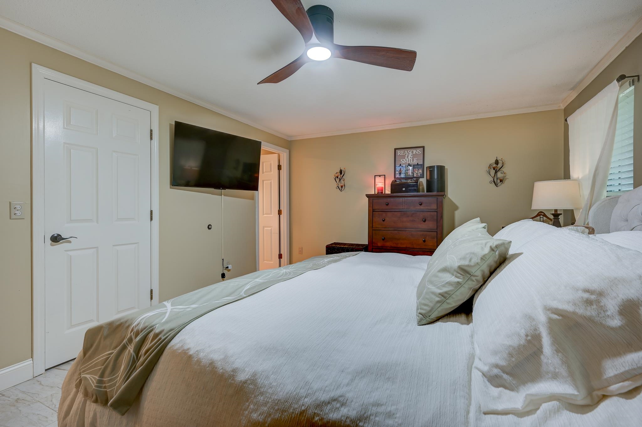 Bedroom with a large white bed, ceiling fan, wall-mounted television, and wooden dresser.