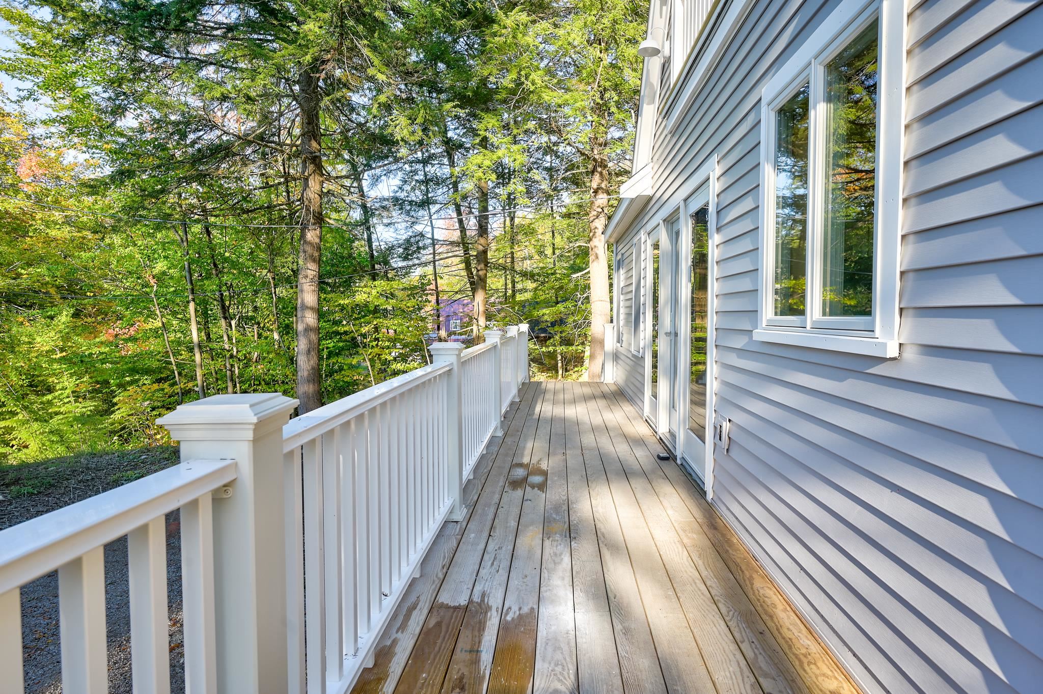 Wooden side deck with white railings beside gray siding, overlooking leafy trees.