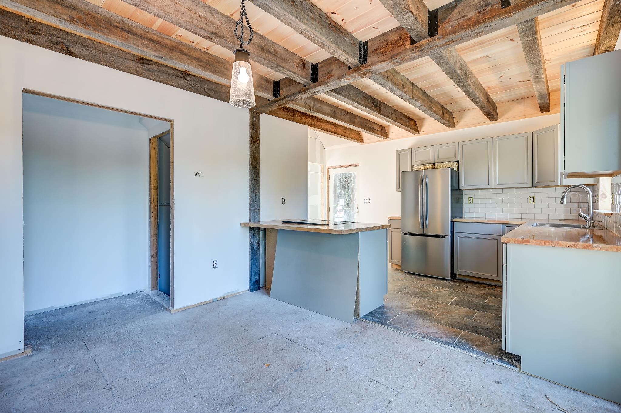 Kitchen with gray cabinets, stainless steel appliances, and exposed timber ceiling beams; adjoining room remains unfinished.